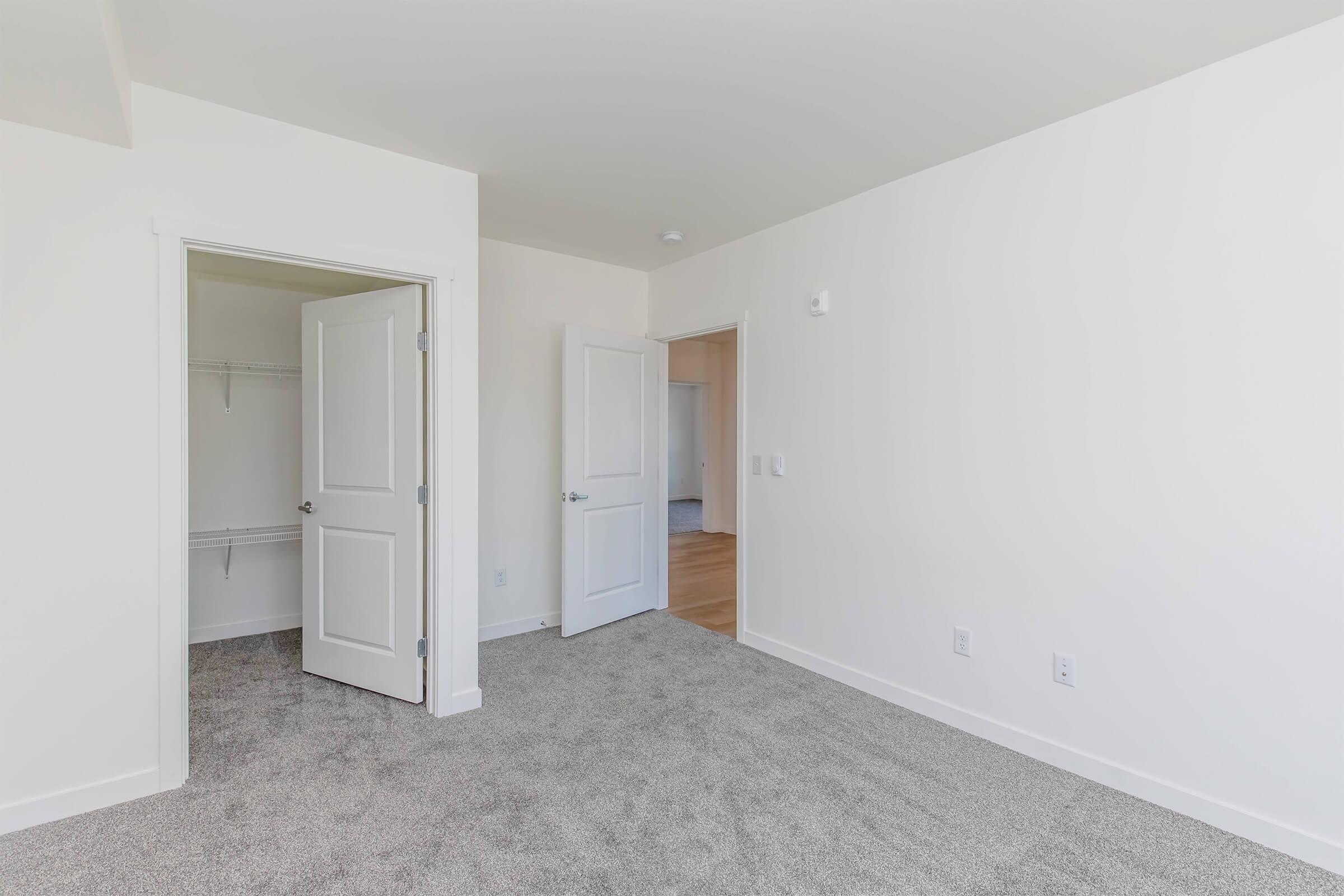 A well-lit, empty bedroom featuring light-colored walls and soft gray carpet. The room has two open doors, one leading to a closet with a shelf. The other door opens to a connected living space, visible in the background. The overall atmosphere is spacious and clean.