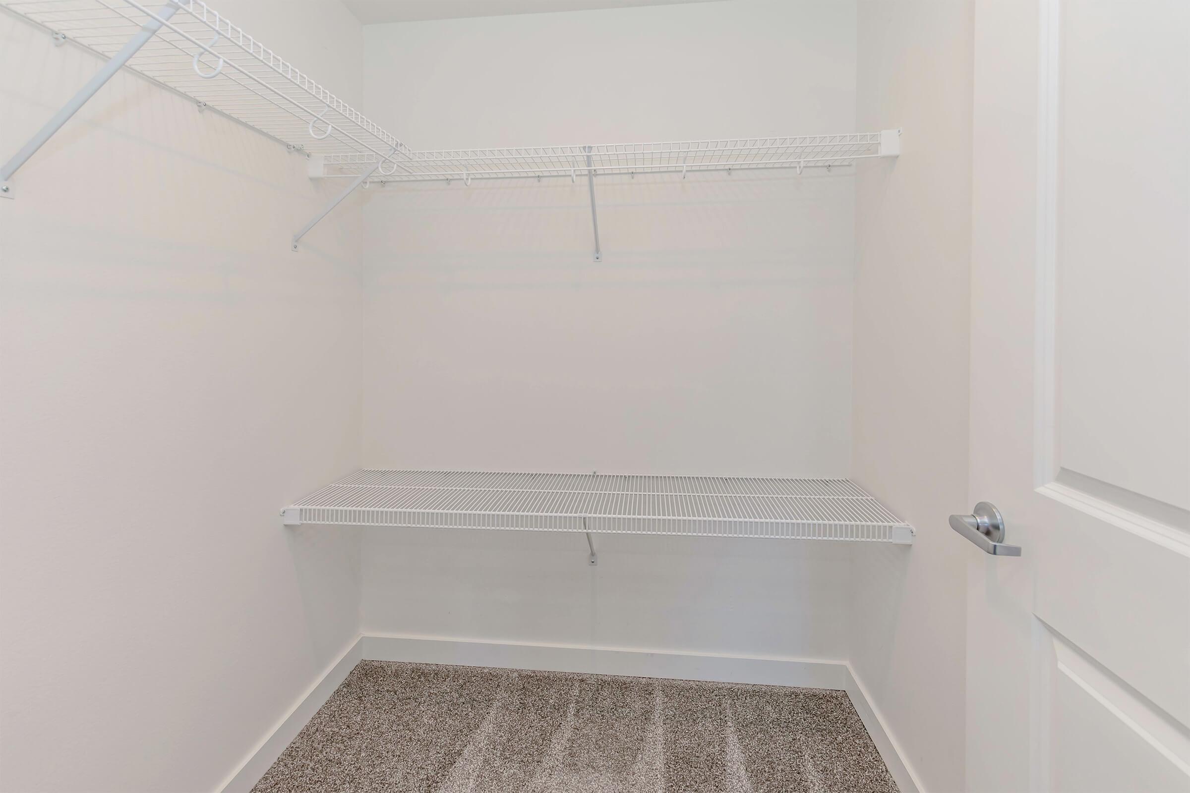 A clean, empty closet featuring white wire shelving on the walls and a wire shelf above a carpeted floor. The room has plain white walls and an open door, suggesting a spacious and minimalistic storage area.