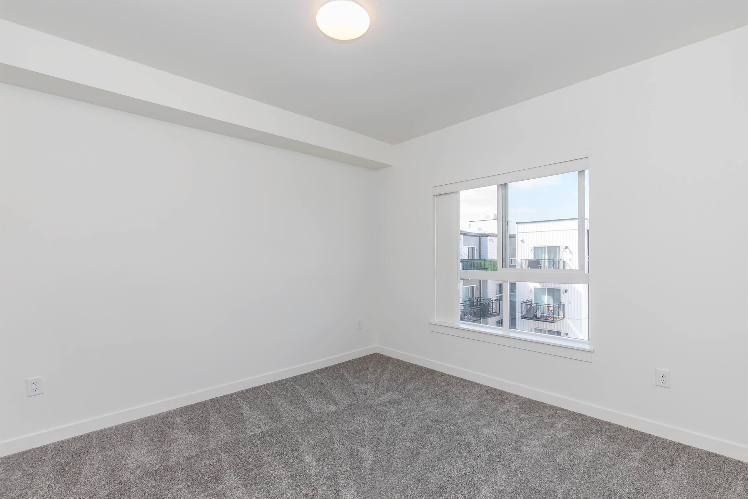 A bright, empty room with light gray carpet and a white wall. A large window with several panes lets in natural light, offering a view of an outdoor balcony area. The ceiling has a round light fixture, and the space has a clean, modern feel, making it suitable for various uses.