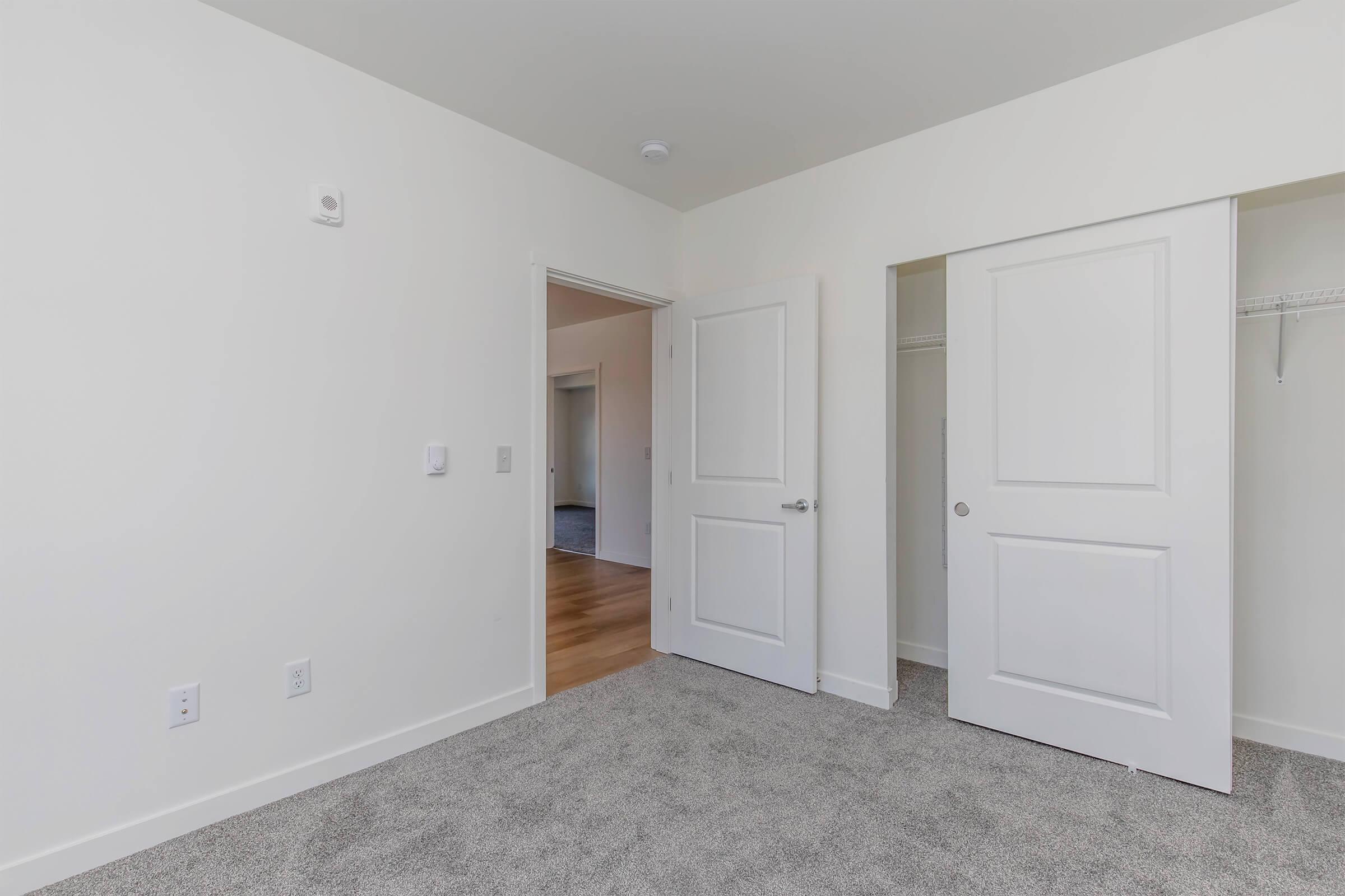 A well-lit room with light-colored walls and carpeted flooring. There is an open door leading to a closet on one side and another door that appears to lead to a different room. The space is uncluttered and features minimal decor, emphasizing a clean and modern look.