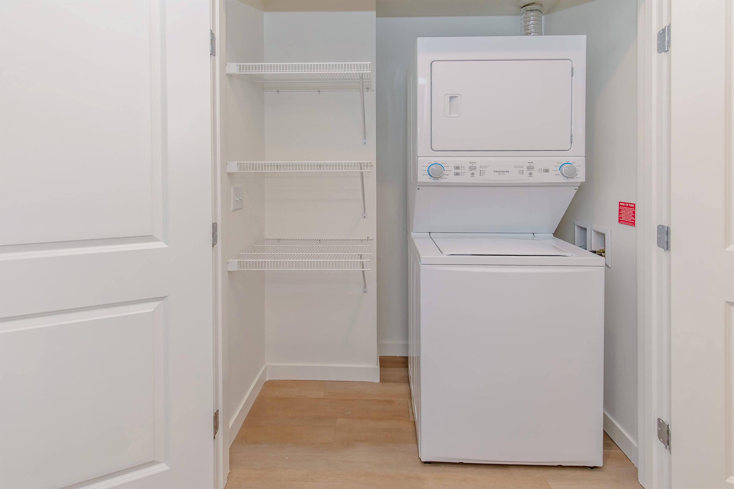 A laundry room featuring a stacked washer and dryer unit, with white shelving on the left side. The walls are painted light, and there is a door leading outside of the room, with a wooden floor.