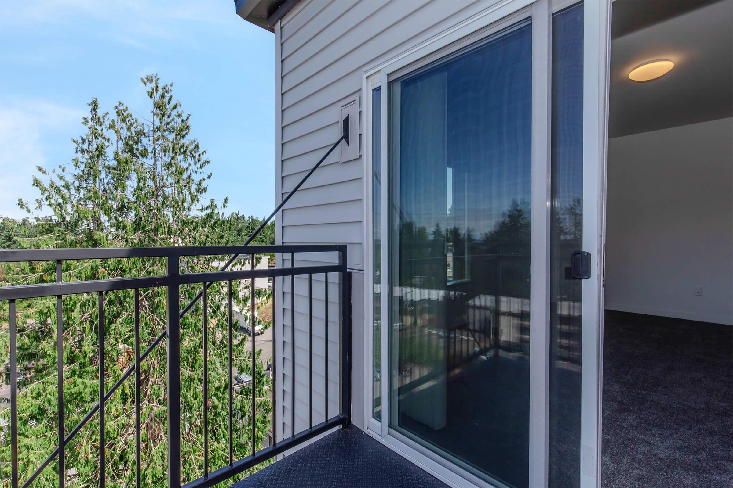 A balcony with a black metal railing, showing an open glass door that leads to an interior space. In the background, trees can be seen, and a bright sky is visible. The interior appears to have carpeted flooring and a ceiling light.