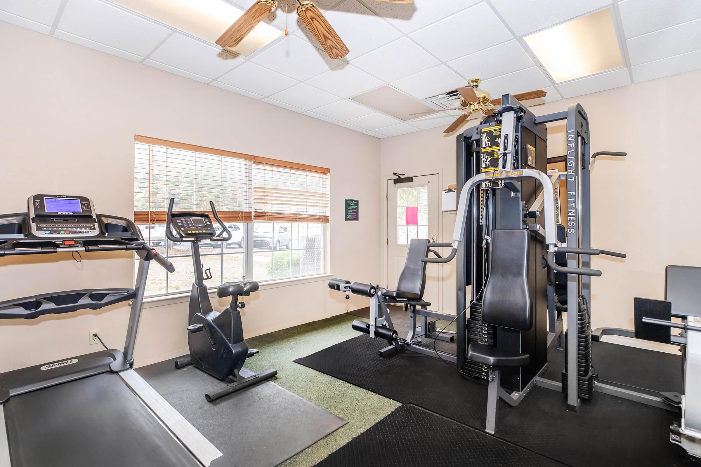 A well-equipped gym setting featuring a treadmill, an exercise bike, and a multi-station weight machine. The room has natural light from windows, a ceiling fan, and rubber flooring.