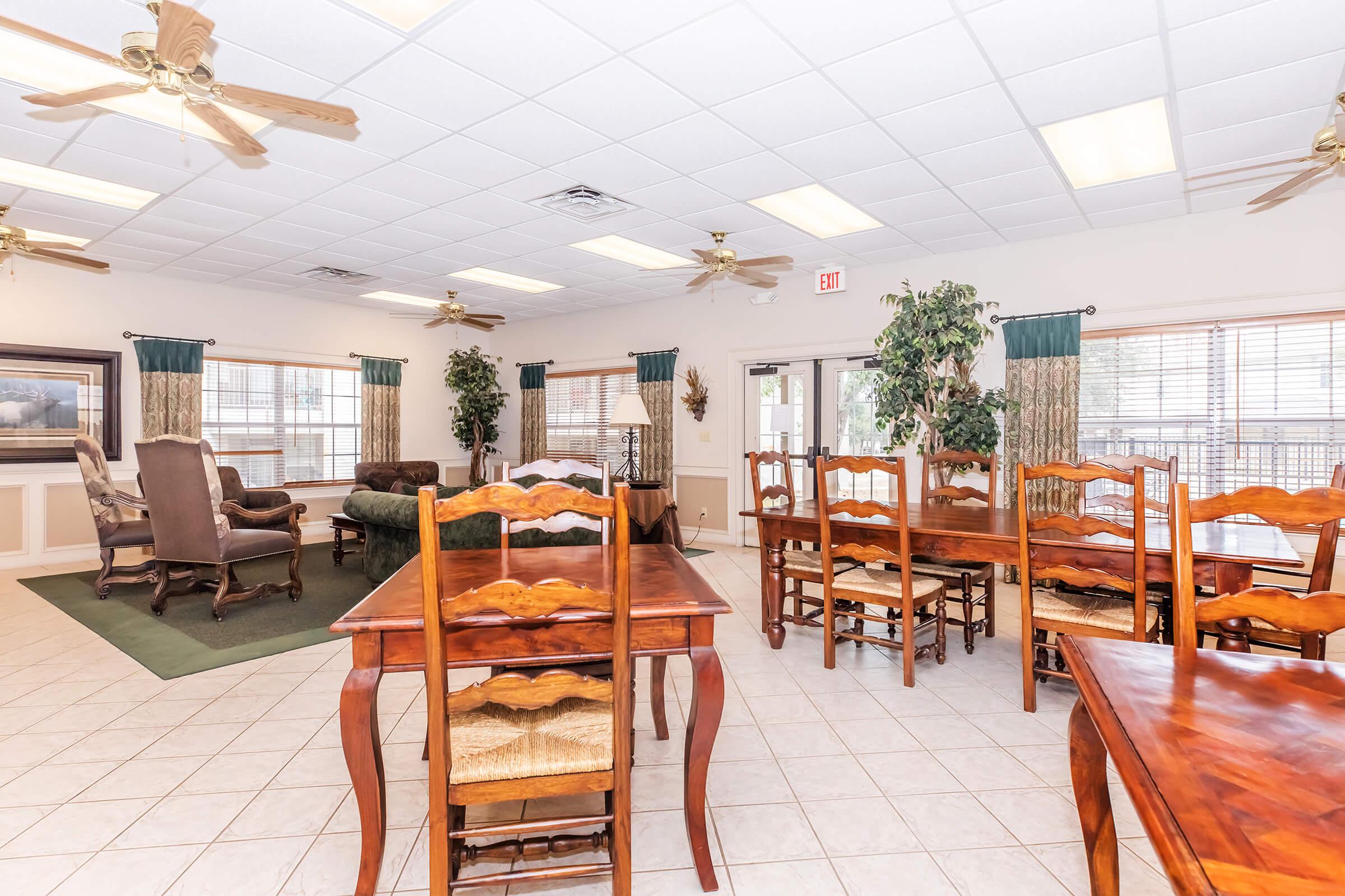 A spacious interior room featuring several wooden dining tables and chairs, with a cozy seating area including a green sofa and armchairs. The room is well-lit with ceiling fans and large windows adorned with curtains. Potted plants are placed strategically around the room, creating a welcoming atmosphere.