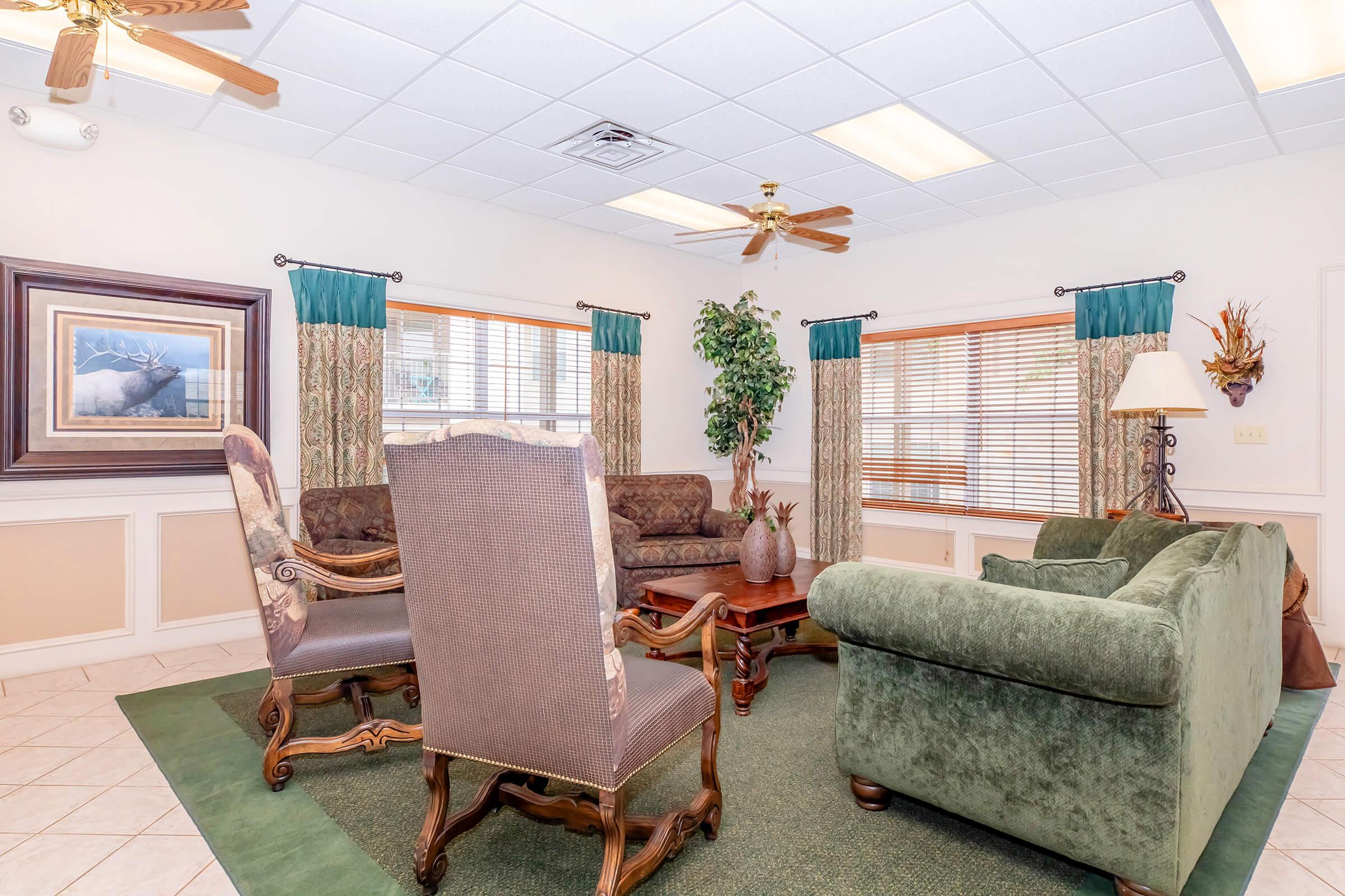 A cozy living area featuring a green upholstered sofa, two armchairs, and a wooden coffee table. The room has patterned curtains, a potted plant, and ceiling fans, with light colored walls and tiled flooring. Natural light comes through the windows, creating a welcoming atmosphere.