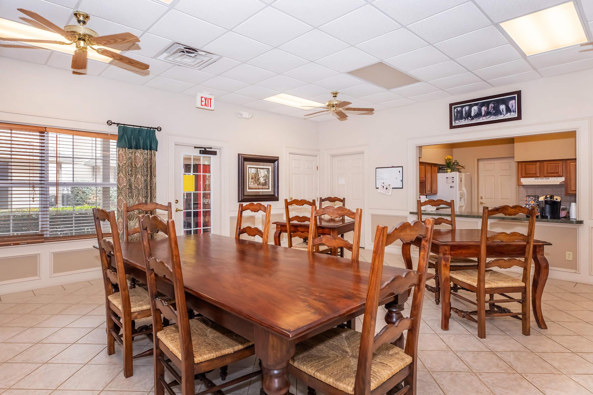 A spacious dining area featuring a large wooden table surrounded by several wooden chairs. The room has a warm, inviting atmosphere with ceiling fans, tile flooring, and natural light coming through windows. A kitchenette is visible in the background, along with a framed image on the wall.