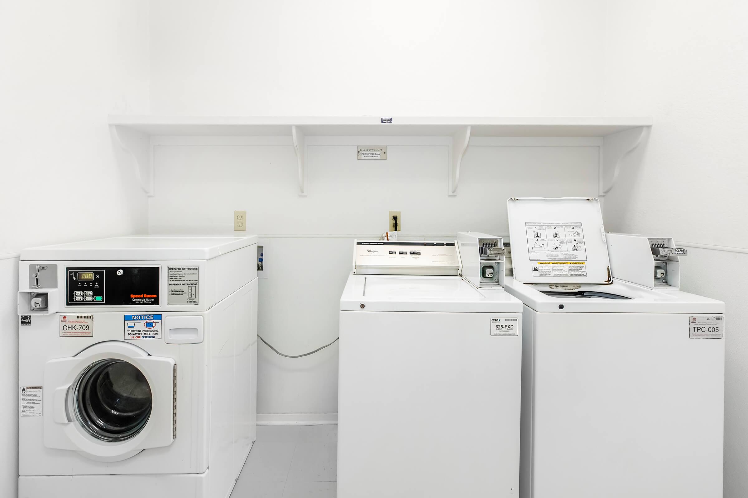 A bright laundry room featuring two washing machines and a dryer. The machines are white and placed against a plain white wall, with a shelf above for storage.