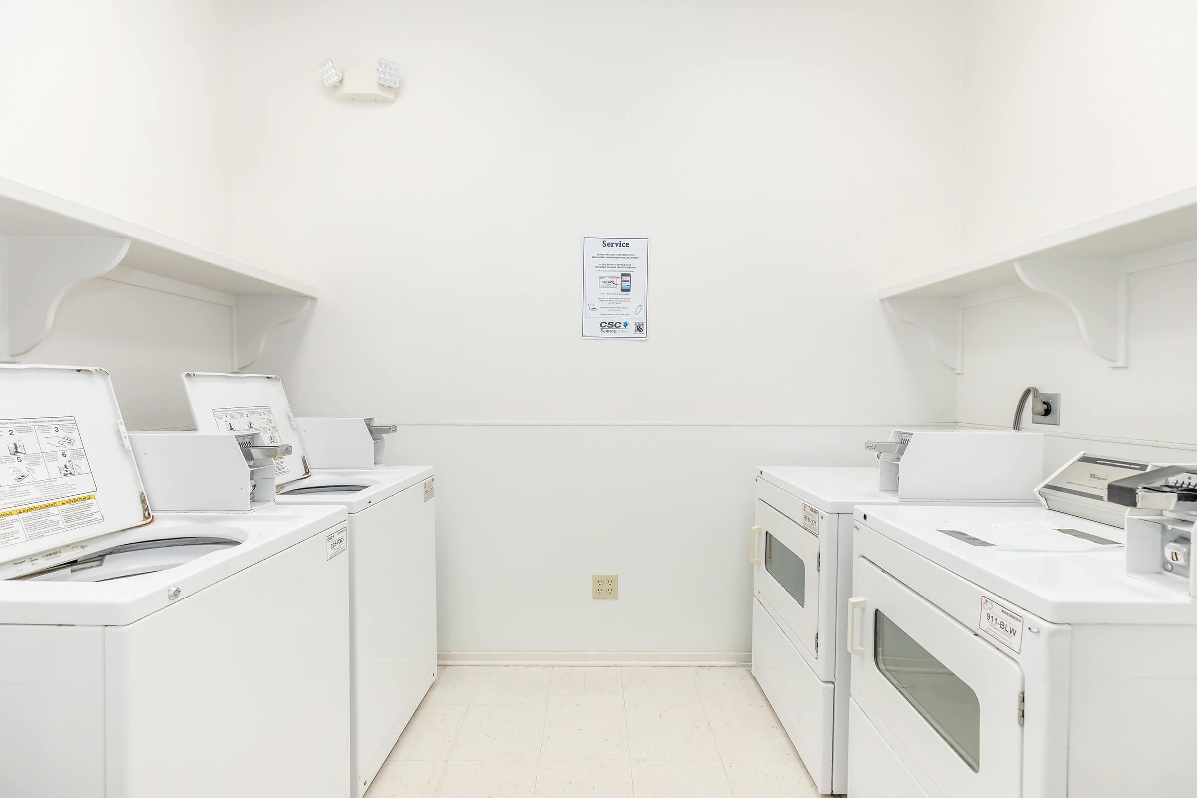 A clean and well-lit laundry room featuring several white washers and dryers lined up against the walls, with a notice or service information posted on the wall. The space has a minimalist design with light-colored walls and a tidy floor.