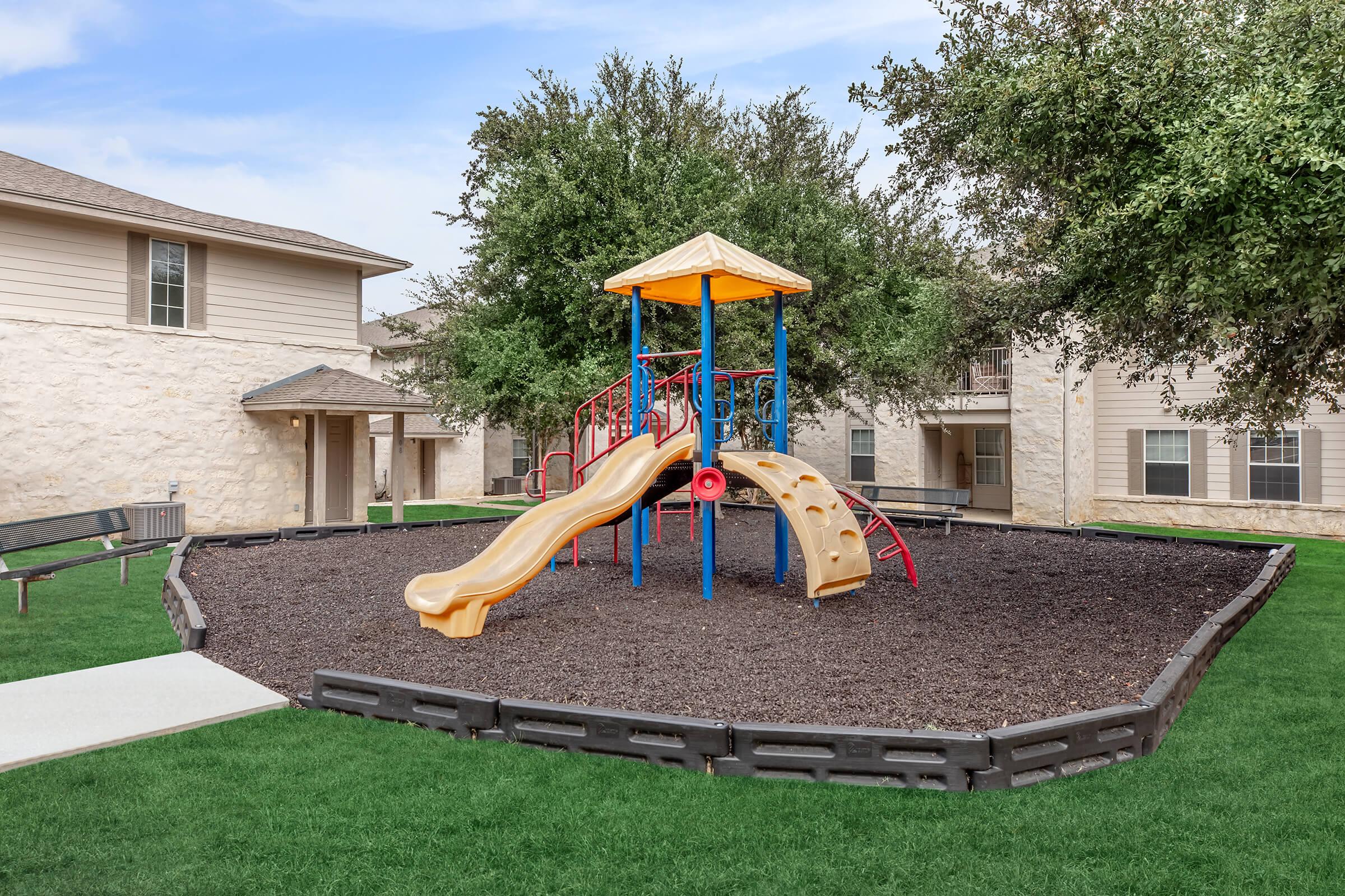 Playground area featuring a colorful slide and climbing structure surrounded by grassy space and trees, set between two residential buildings.