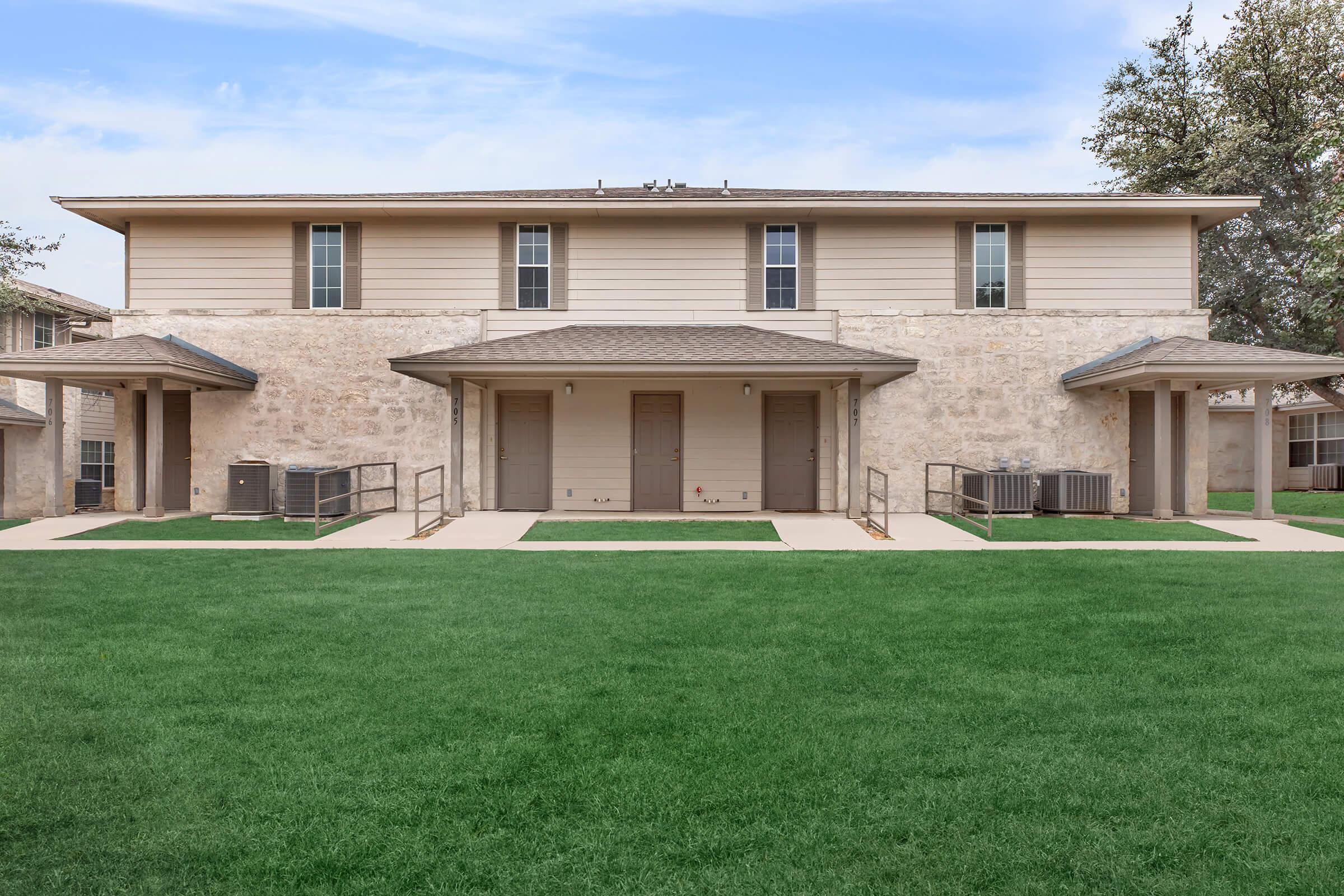 A two-story residential building made of stone and siding, with large windows and two covered entrances. The building is surrounded by a well-maintained green lawn.