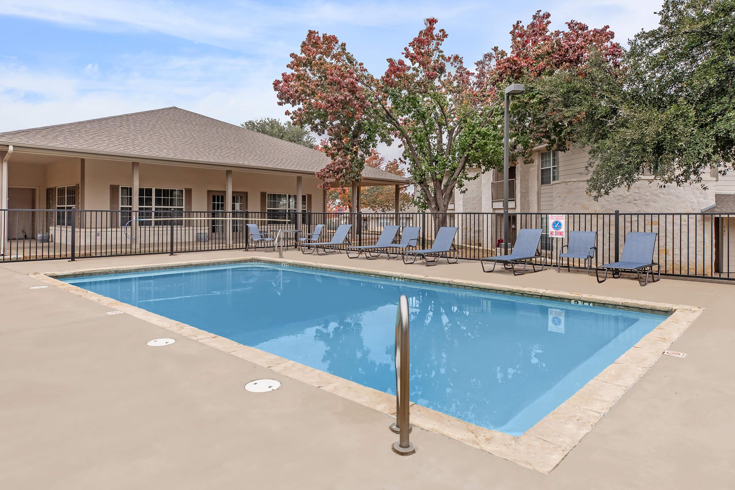 A clear swimming pool surrounded by lounge chairs, with a residential building and trees in the background. The pool area features a light-colored concrete deck and a fence around the pool.