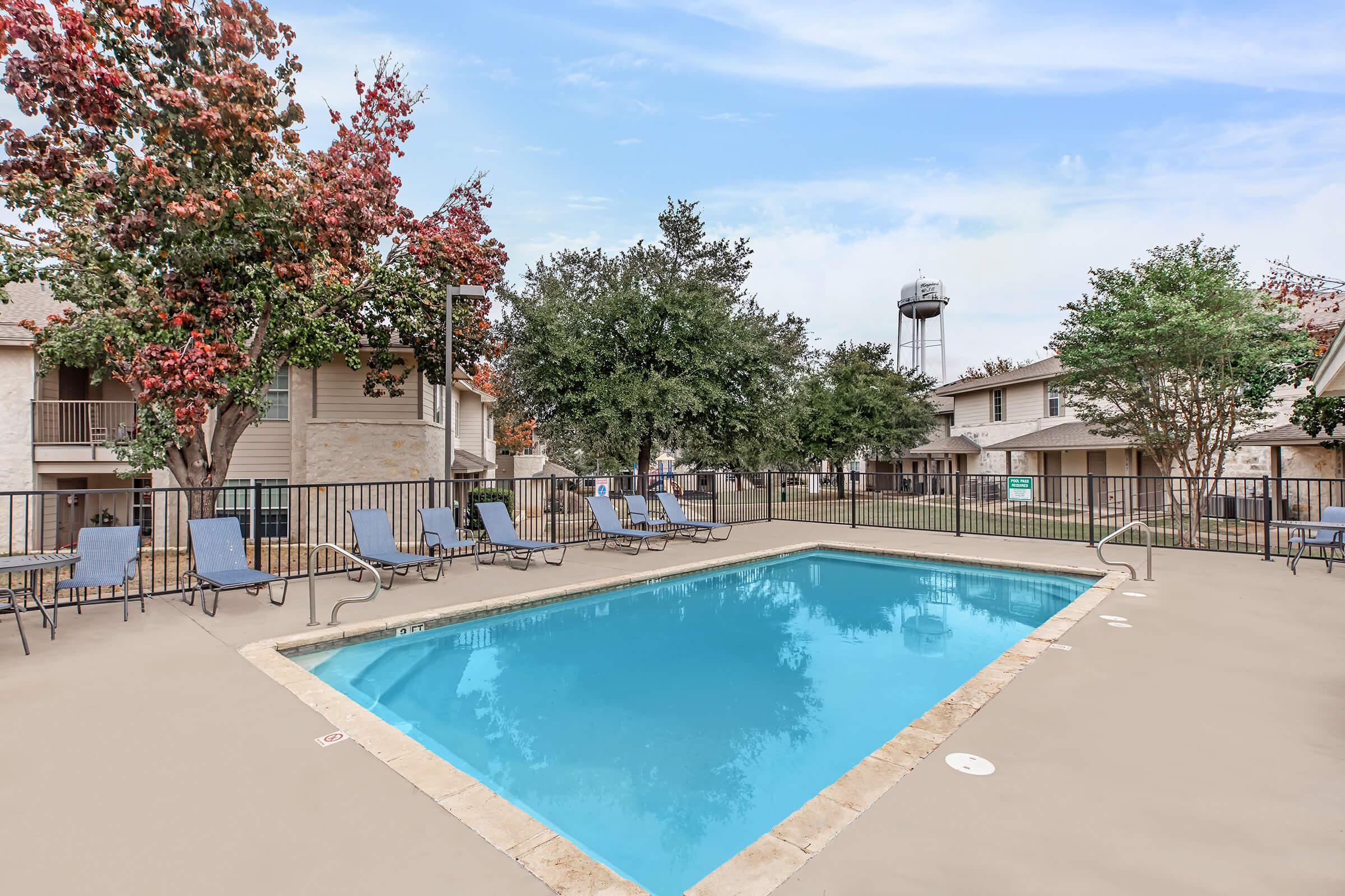 A clear swimming pool surrounded by lounge chairs, with trees and apartment buildings in the background, and a water tower visible in the distance. The scene is set on a sunny day with a blue sky.