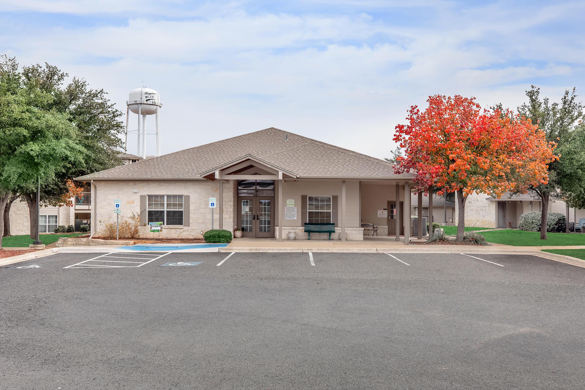 A single-story building with a large entrance, surrounded by trees, one of which has bright red foliage. A water tower is visible in the background. There is a parking area in front with several parking spaces.