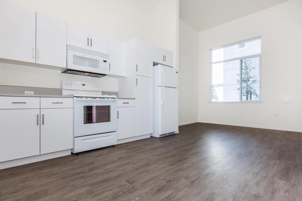 Modern kitchen featuring white cabinetry, a stainless steel microwave above a white stove, and a tall white refrigerator. The room has a high ceiling with large windows allowing natural light to illuminate the space. The floor is finished with a light wood laminate for a contemporary look.
