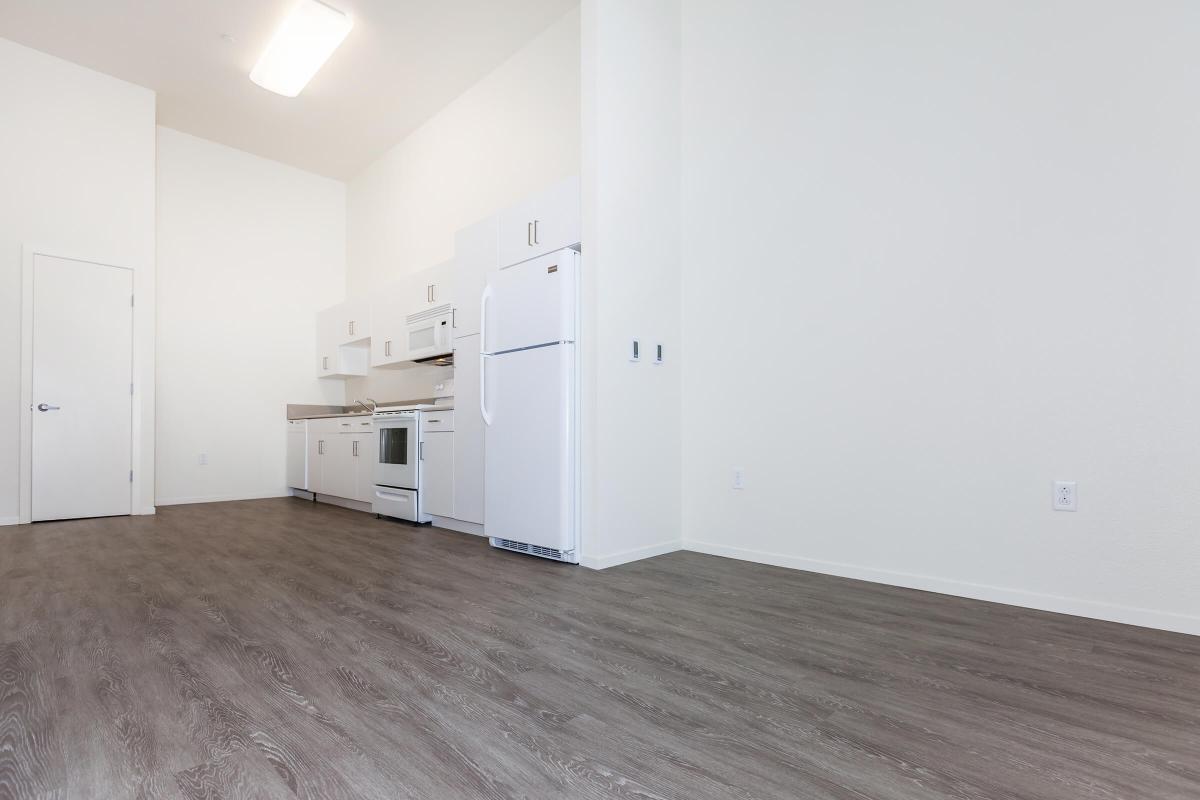 A bright, modern kitchen area featuring white cabinets and appliances, including a refrigerator and oven. The floor is covered with light-colored laminate, complemented by white walls and a high ceiling, creating an open and spacious feel.