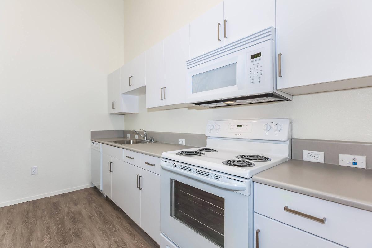 A modern kitchen featuring white cabinets, a microwave above an electric stove, and a sink. The countertop is light gray, and the flooring is a wood-like laminate. The kitchen has a spacious and airy feel, with neutral-colored walls and ample storage.