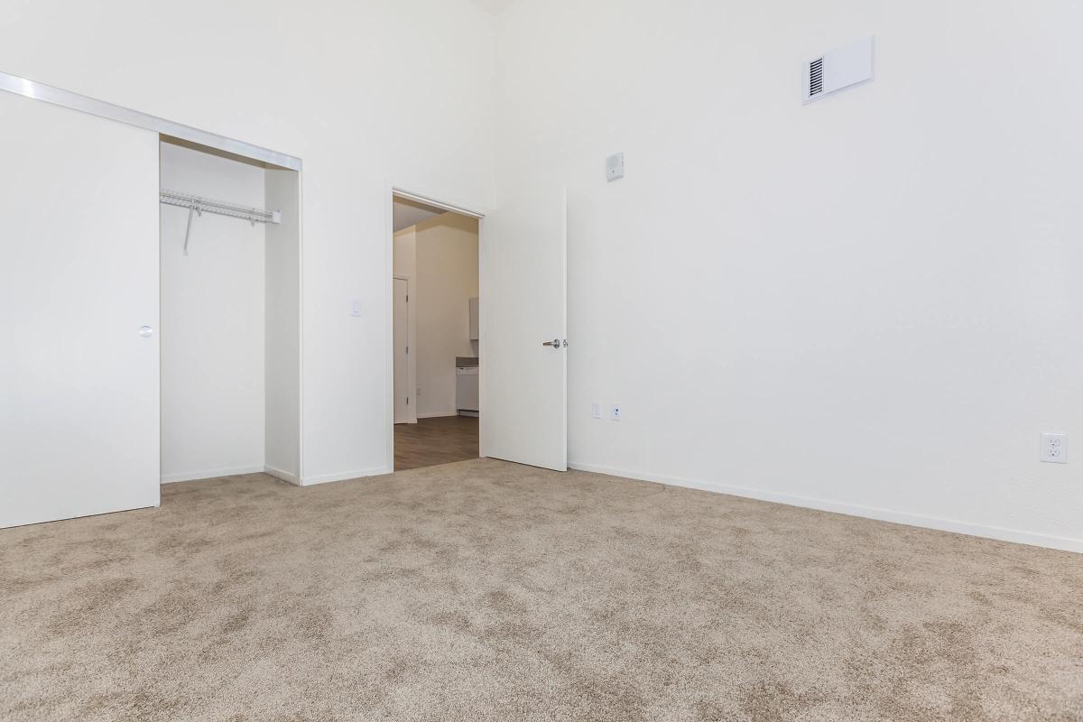 A spacious, empty room with beige carpet flooring, featuring two open white doors. One door leads to a closet, and the other opens to a bathroom area out of view. The walls are painted white, and there is a vent on the wall. Natural light brightens the space, creating an inviting atmosphere.