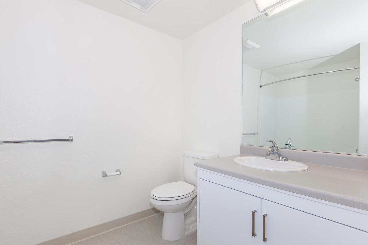 A clean, minimalist bathroom featuring a toilet, a sink with a countertop, and a large mirror. The walls are white, and the flooring is light-colored. A towel bar is mounted on the wall, and there is a glass shower enclosure in the background, indicating a modern design.