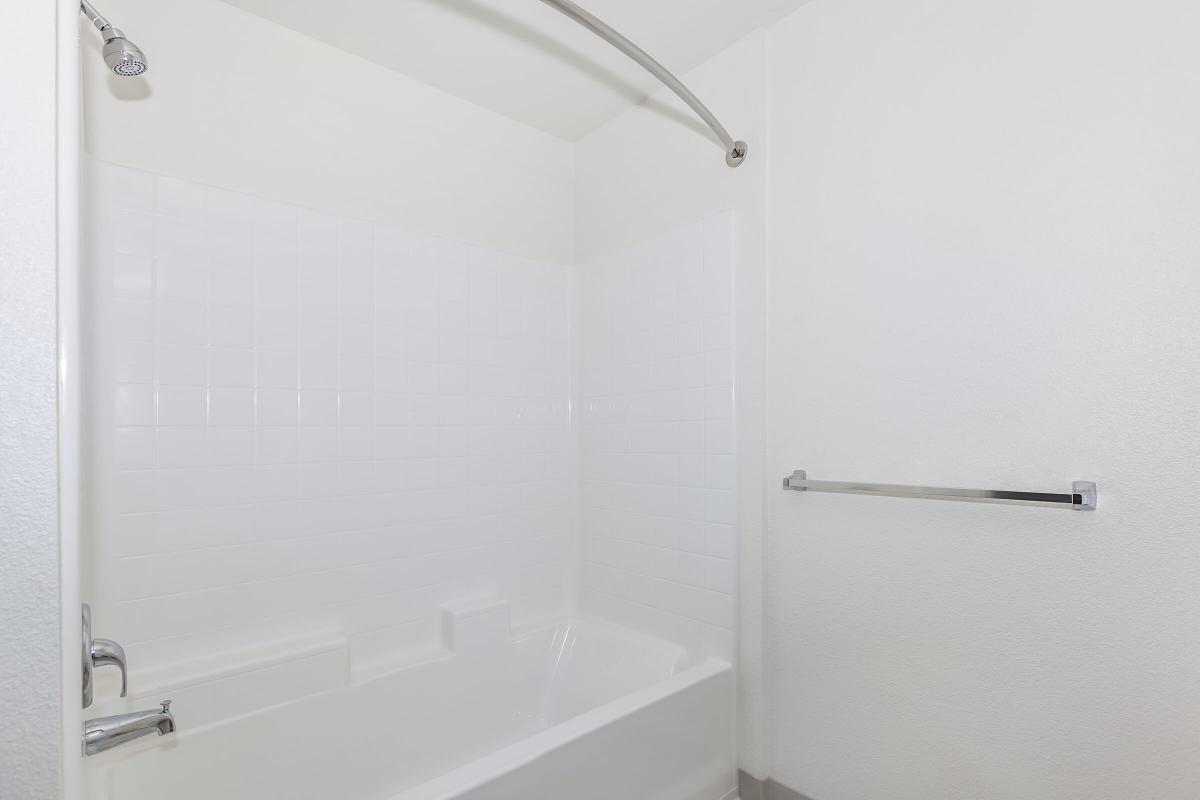 A clean and minimalistic bathroom with a bathtub and shower combination. The walls are tiled in white, and there is a chrome towel bar installed on the wall. The overall design is simple and bright, creating an airy atmosphere.