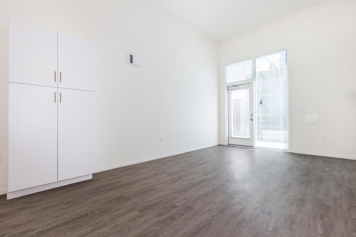 A minimalistic interior space featuring light-colored walls and a wooden floor. There is a tall white cabinet against the left wall and a large glass door on the right leading to an outdoor area. The room is well-lit, creating an open and airy atmosphere.