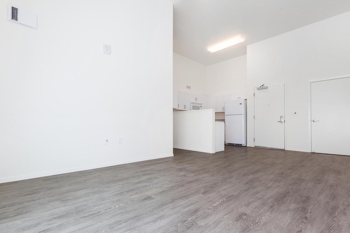 A spacious, modern interior featuring light-colored walls and a wooden floor. The image shows an open layout with a kitchen area in the background, including a refrigerator and a closed door on the right. Natural light illuminates the space, creating a welcoming atmosphere.