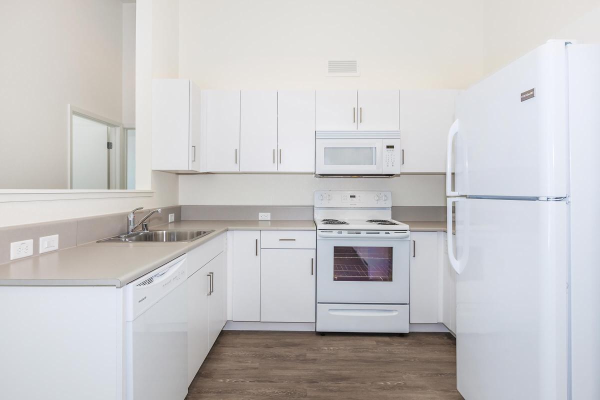 Bright and modern kitchen featuring white cabinets, stainless steel sink, white appliances including a microwave and oven, and a large refrigerator. The countertops are a light color, and the flooring appears to be wood. The space is well-lit and organized, creating a clean and inviting atmosphere.