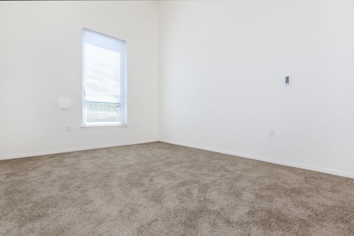 A spacious empty room with light-colored walls and carpeting. There is a window with vertical blinds allowing natural light to enter, and a small wall-mounted device visible on one side. The room is minimalistic and uncluttered, creating a bright and open atmosphere.