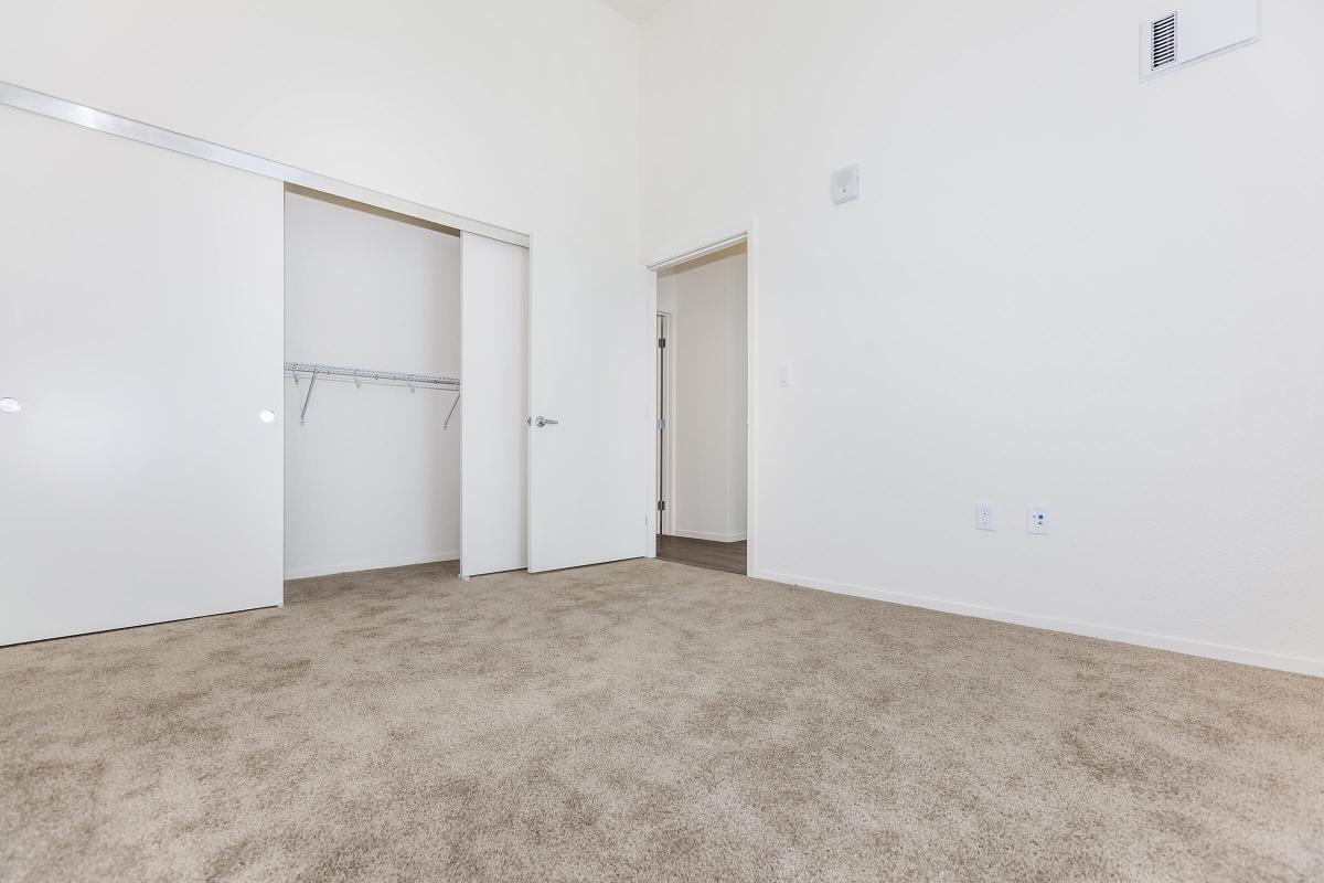 A spacious room with light beige carpet, featuring white walls and two open doors. One door leads to a closet with a hanging rod, while the other opens to an adjacent area. The room has a bright and airy atmosphere, with minimal furnishings.