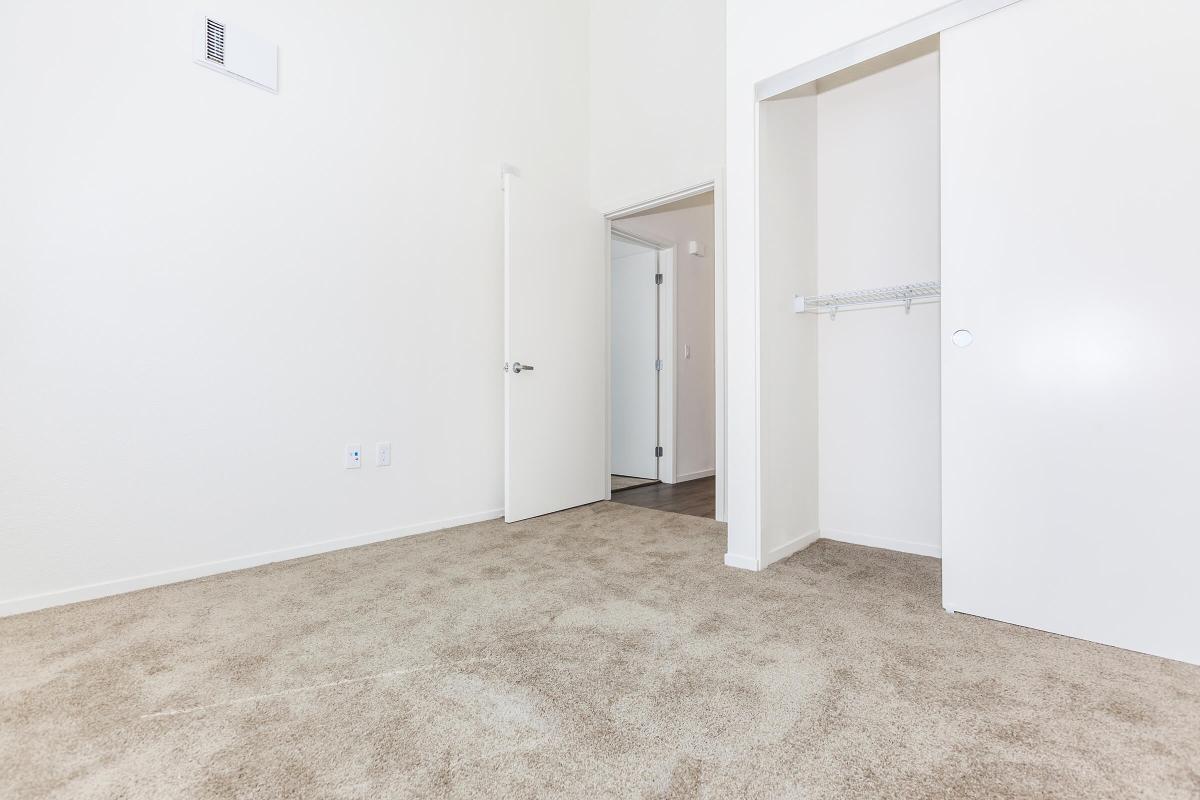 A lightly colored room with beige carpet and white walls, featuring an open doorway leading to another room. There is a closet with sliding doors on the right and a small wall-mounted shelf. Natural light brightens the space, giving it a clean and airy feel.