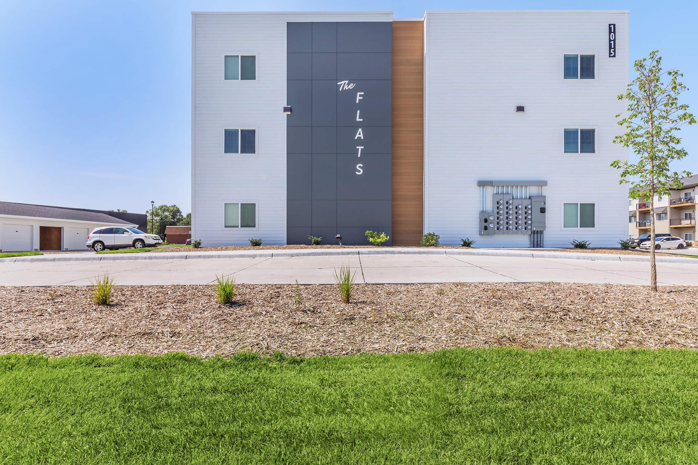 Modern apartment building with a sleek exterior featuring a mix of white siding and gray accents. The building is three stories tall, displaying the name "The Flats" prominently. Well-kept landscaping with green grass and small plants is visible in the foreground, along with parking spaces for vehicles.