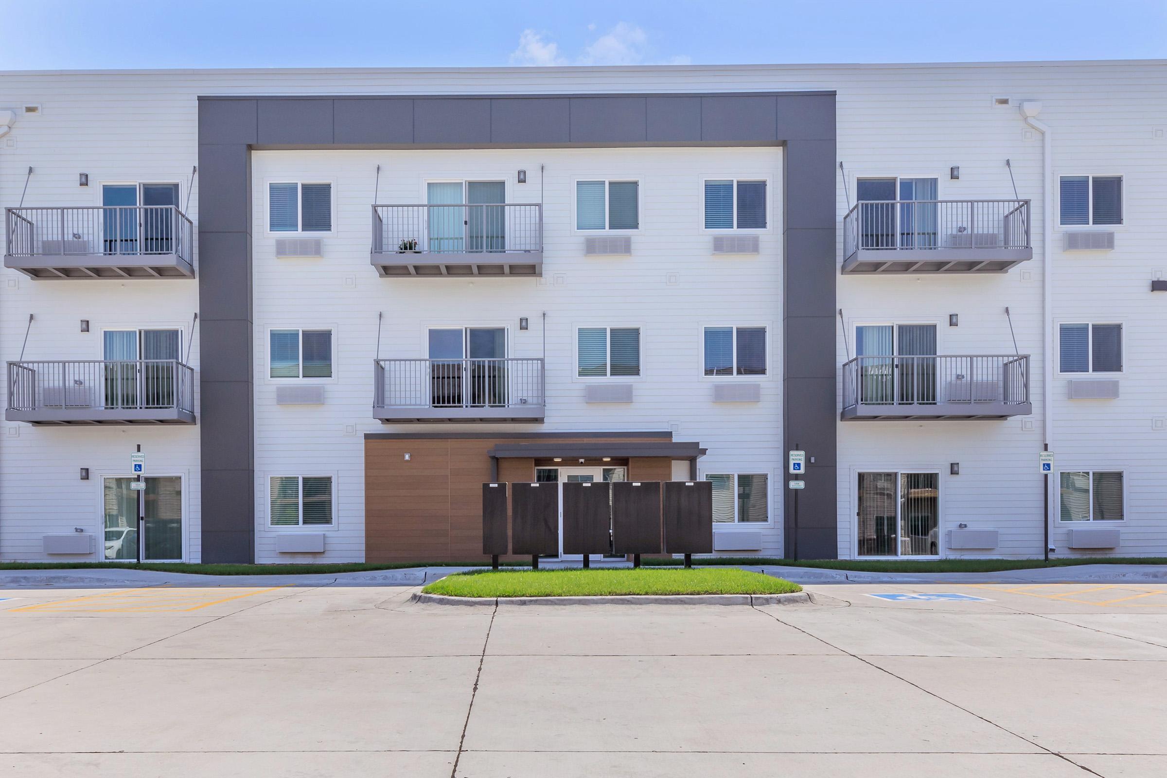 Three-story modern apartment building with a white exterior and gray accents. Each floor features multiple windows and balconies. The entrance is at the center, flanked by landscaped grass areas. The surrounding area includes a paved driveway and parking spaces in front of the building.