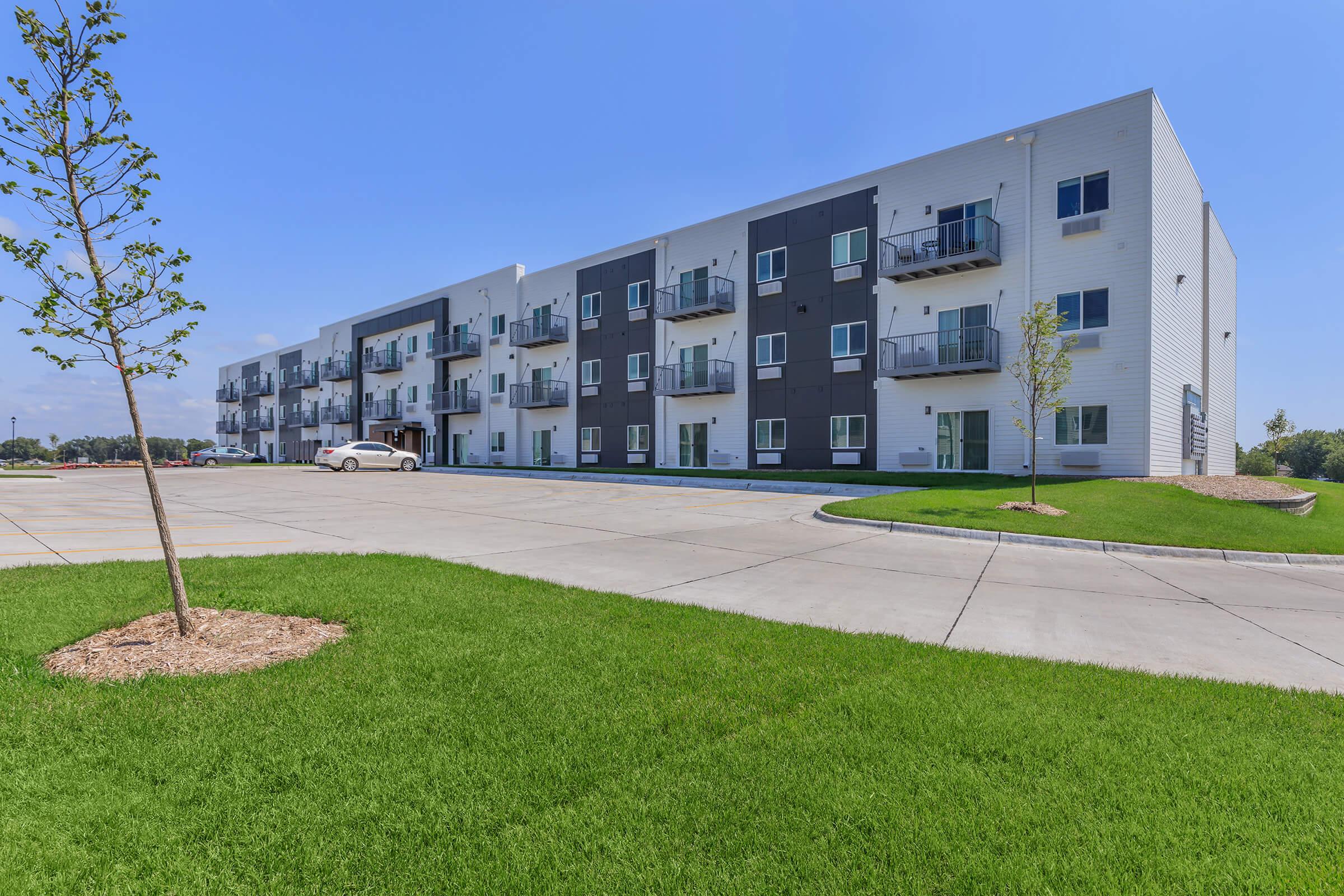 Modern residential building with a sleek design, featuring multiple balconies and large windows. The surrounding area has well-maintained green grass and a young tree. A portion of the paved parking lot is visible, along with a parked white car. The sky is clear and bright.