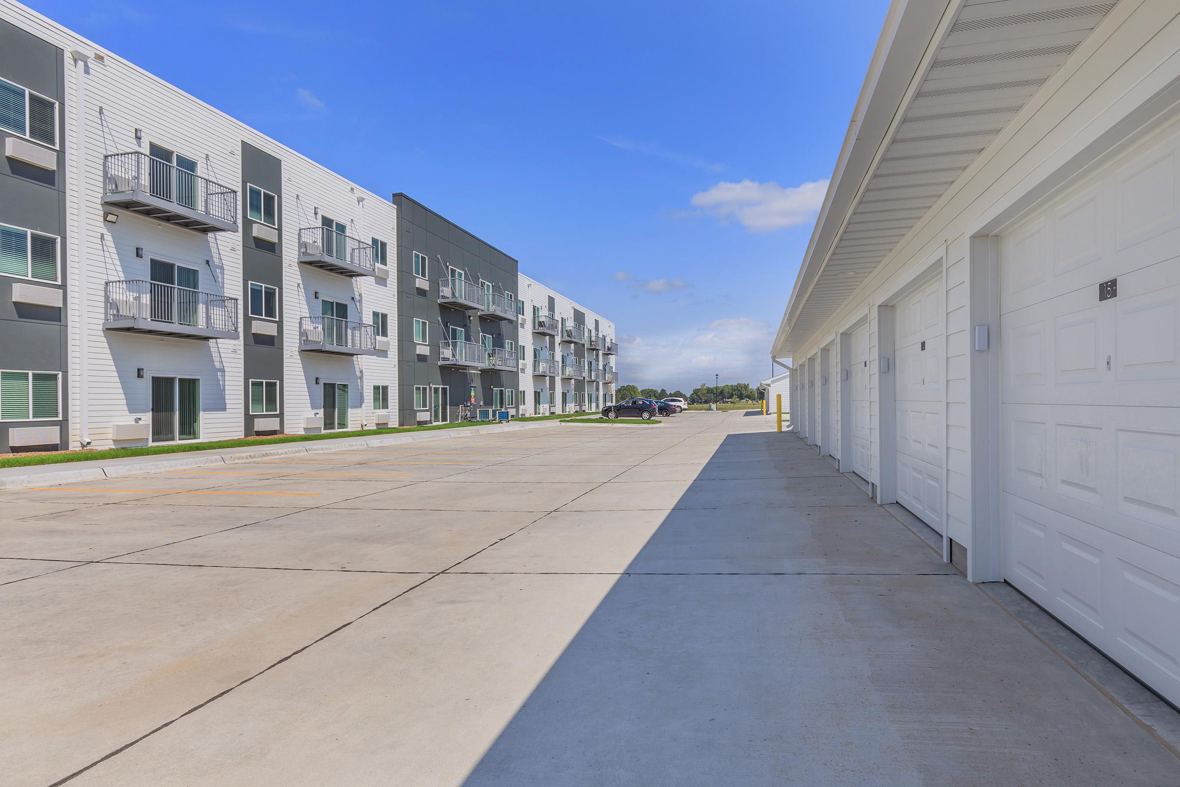 A modern residential complex featuring two buildings with multiple apartments, balconies, and large windows, alongside a row of garage doors. The scene is set under a clear blue sky with some clouds, and a paved parking area extends between the structures.