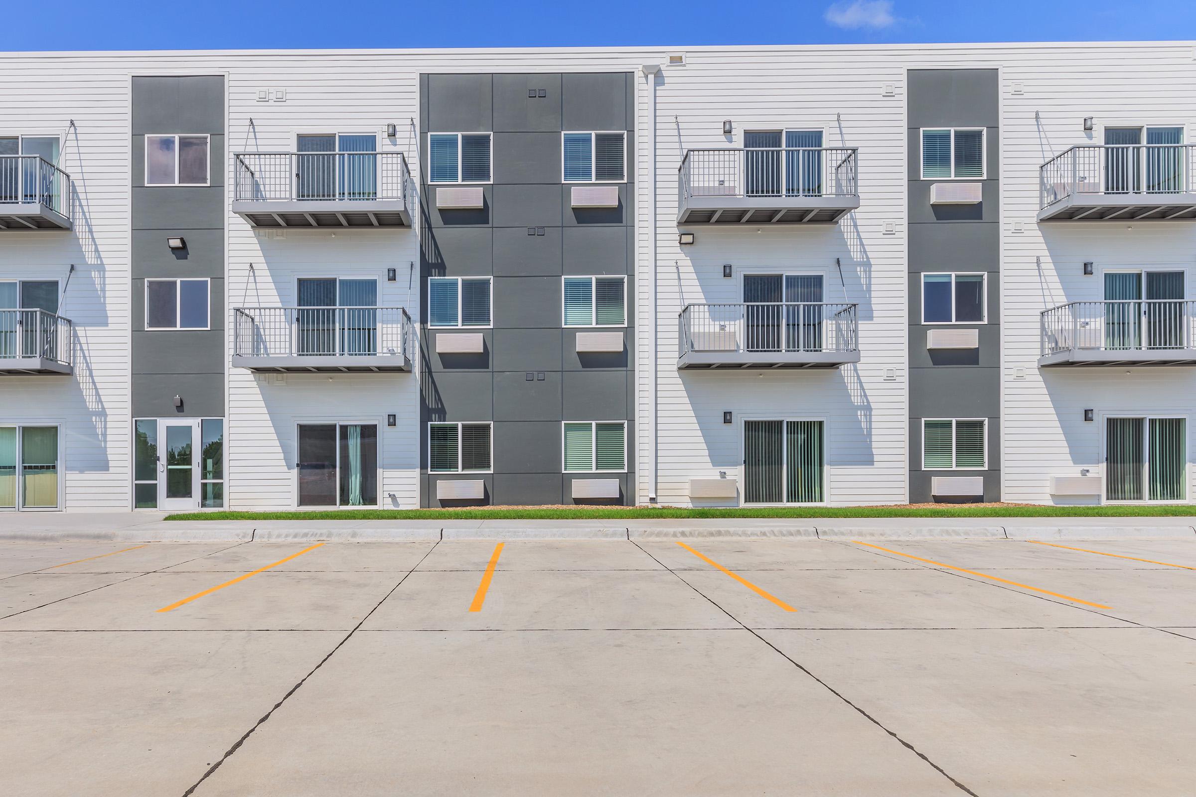 A modern apartment building with a sleek design featuring multiple balconies, large windows, and a mix of white and gray exterior. In the foreground, there is a parking lot with painted lines, and the sky is clear with a few clouds visible.