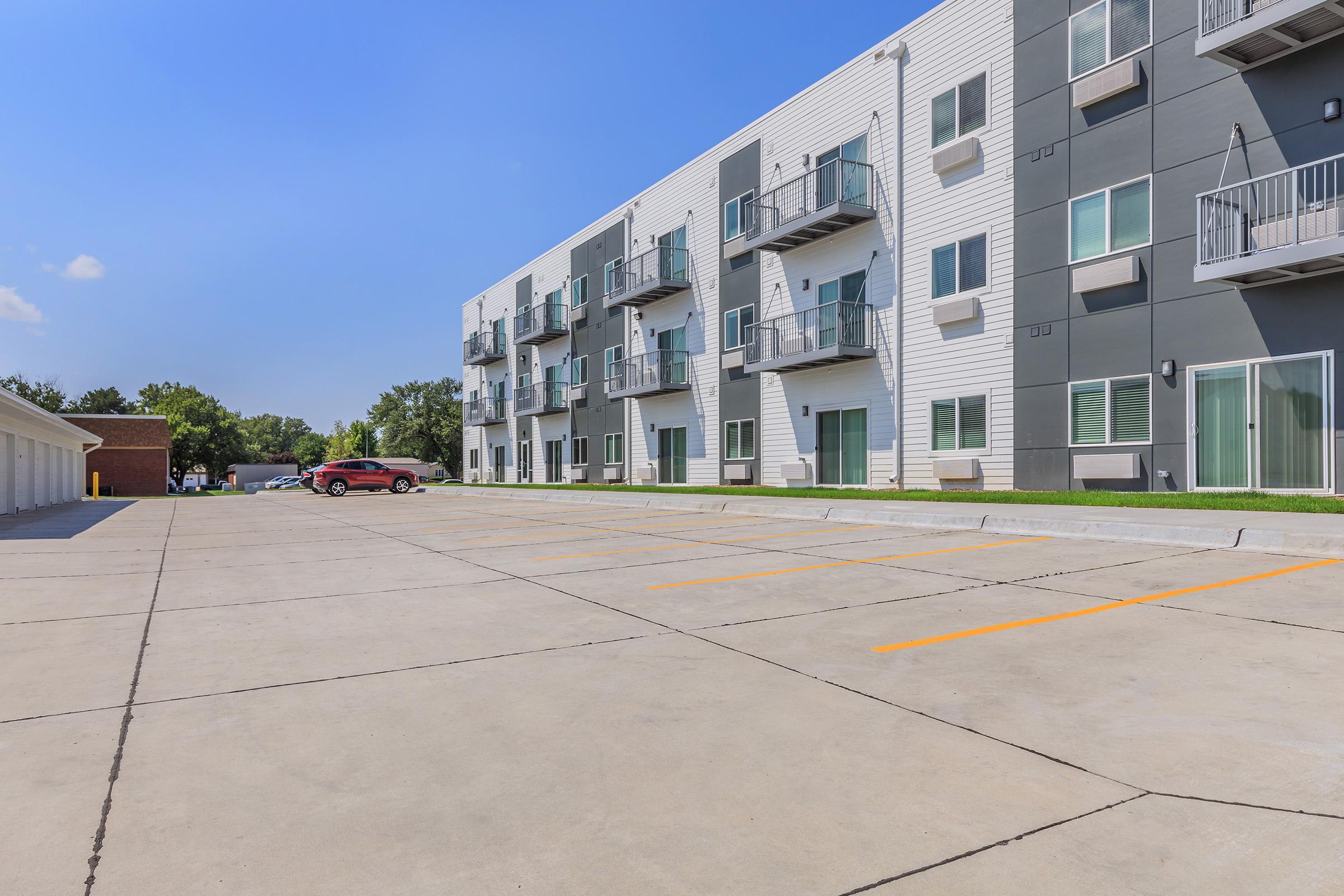 Modern apartment building featuring a mix of light and dark siding, with multiple balconies. A spacious parking lot is in the foreground, partially empty with several marked parking spaces. Clear blue skies above and lush greenery in the background.