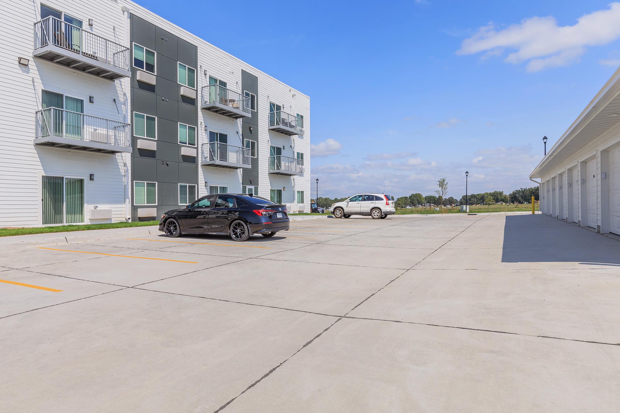 A modern apartment building with a mix of white and gray exterior, featuring a few balconies. In the foreground, there are two parked cars on a concrete surface with clearly marked parking spaces. The background shows an open area with greenery and a blue sky with scattered clouds.