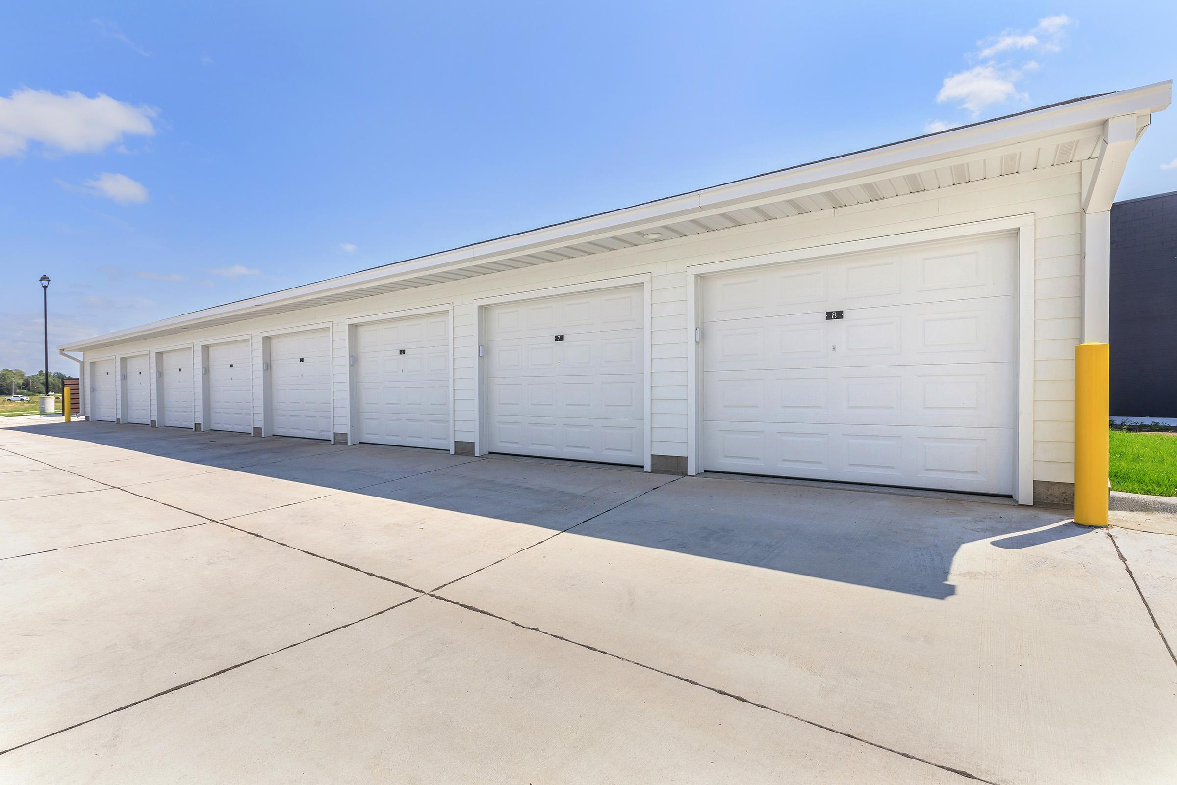 A row of white garage doors lined up side by side in a well-lit outdoor area. The concrete floor is smooth, and the sky is clear with a few clouds. There is a yellow post on the right side of the image, adding a pop of color to the scene.