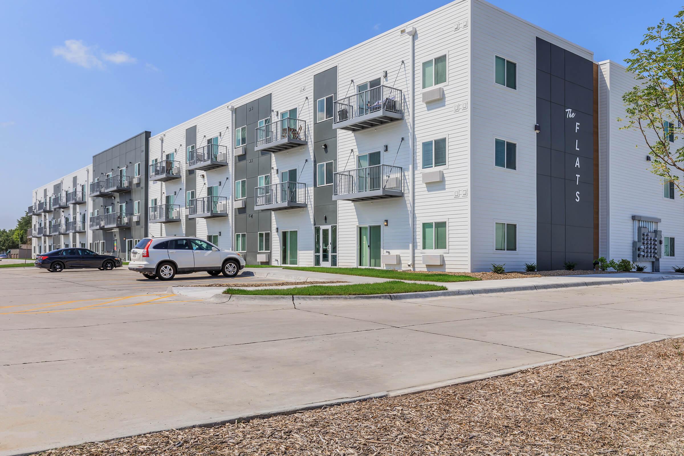 A modern apartment building with a white exterior and gray accents. It features multiple balconies, large windows, and the name "The Flats" displayed prominently on the side. A parking lot in front has a few cars, and the landscaping includes grass and gravel areas under a clear blue sky.