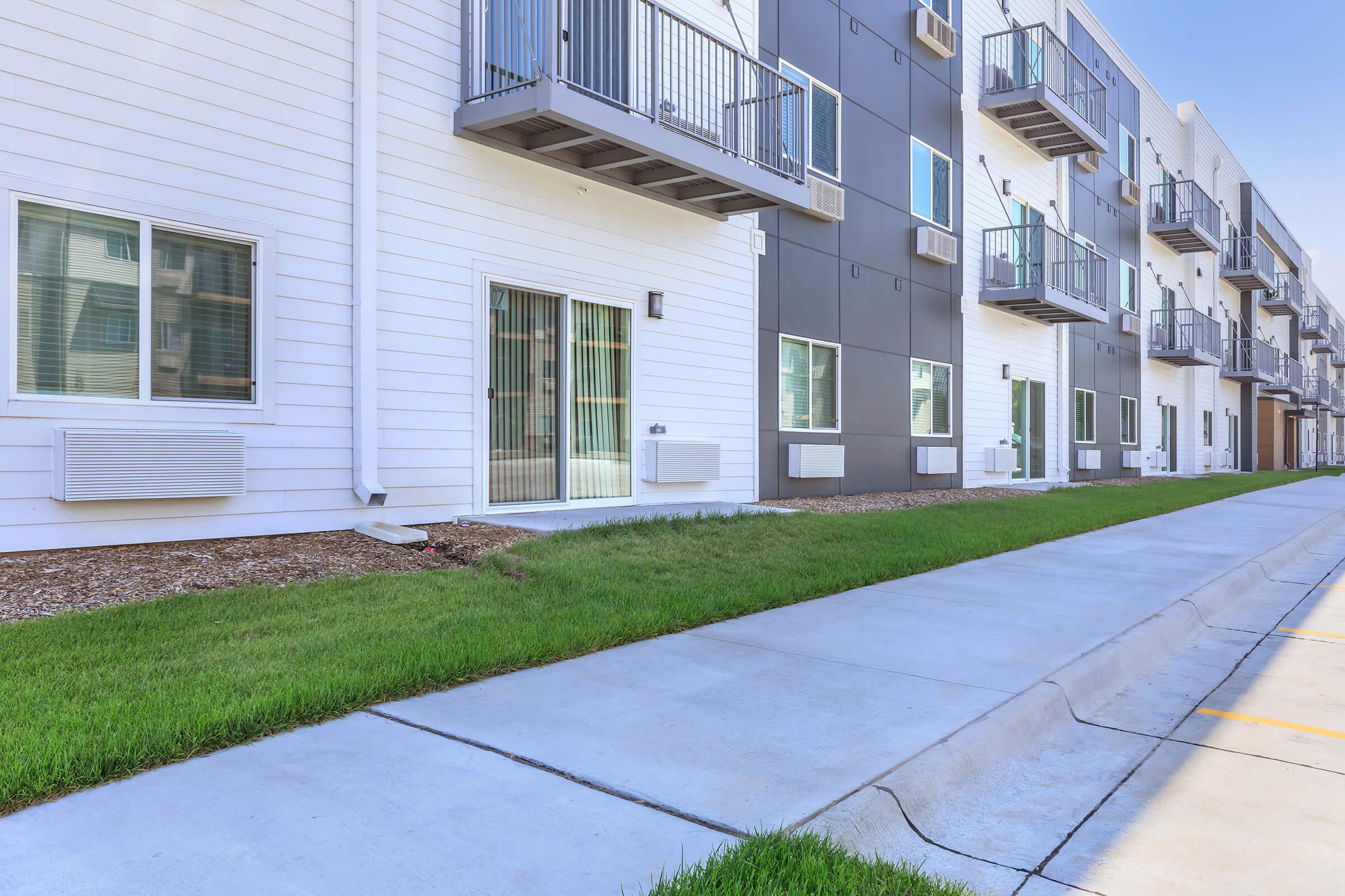 Modern apartment building exterior with multiple units featuring balconies. The area includes a well-maintained lawn and a concrete walkway. Air conditioning units are visible below the windows, adding to the contemporary urban aesthetic.