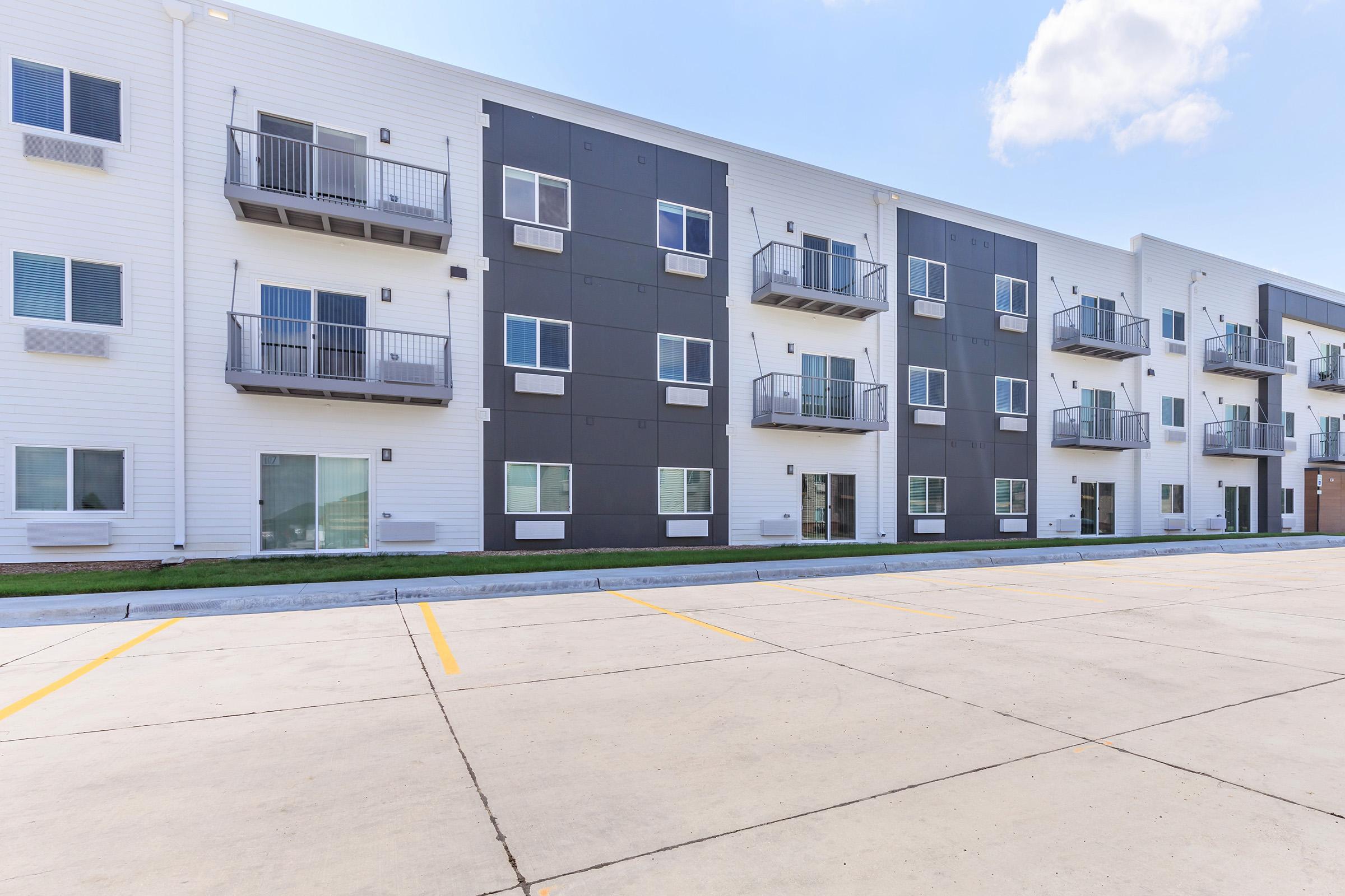 Modern apartment building with a mix of white and dark-colored exterior walls. Features multiple floors with balconies and large windows. A flat, paved parking area is visible in the foreground, and a clear blue sky is present above the building.