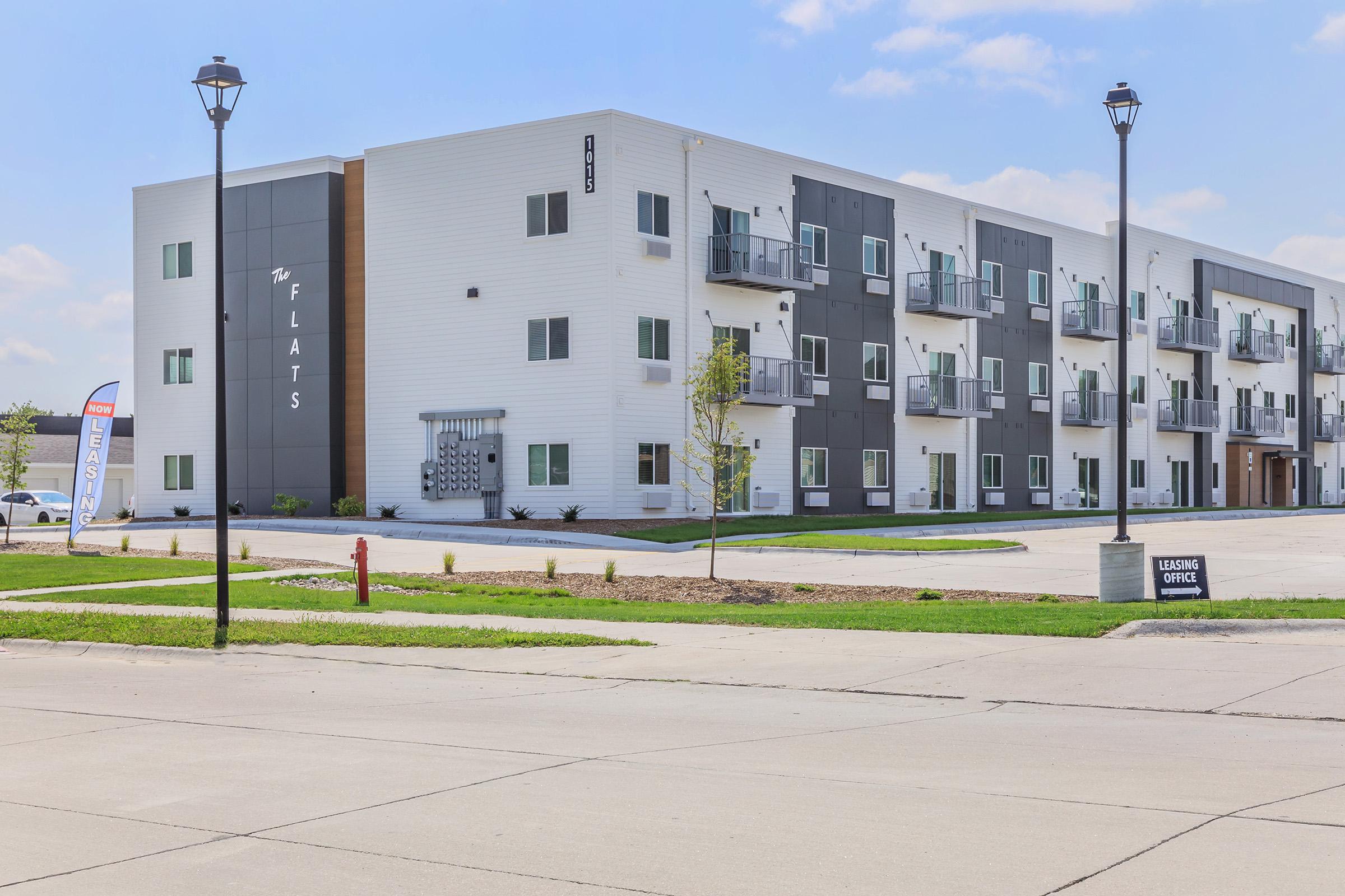 A modern apartment building featuring multiple units with balconies, large windows, and a stylish facade. The building is surrounded by landscaped areas and is situated near a parking lot and street, with a sign indicating a leasing office nearby. The sky is clear with a few clouds.