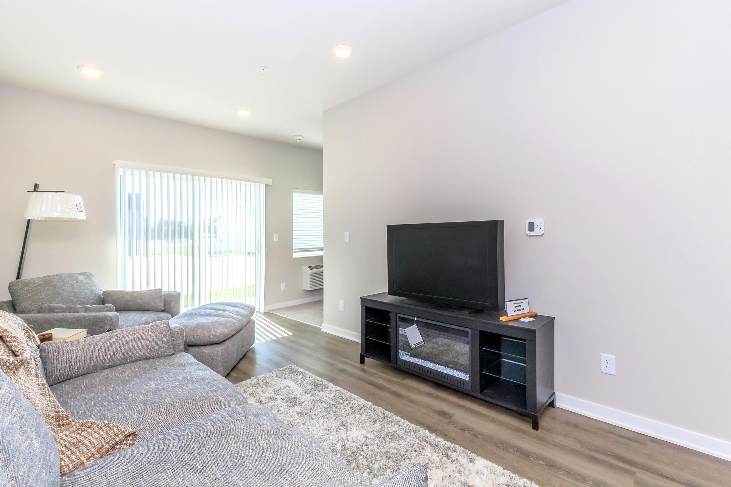 A modern living room featuring a gray sofa and armchair, a black TV stand with a television, and natural light coming through vertical blinds. The walls are painted in a light color, and there is a small table next to the sofa. The space has a clean and contemporary design.