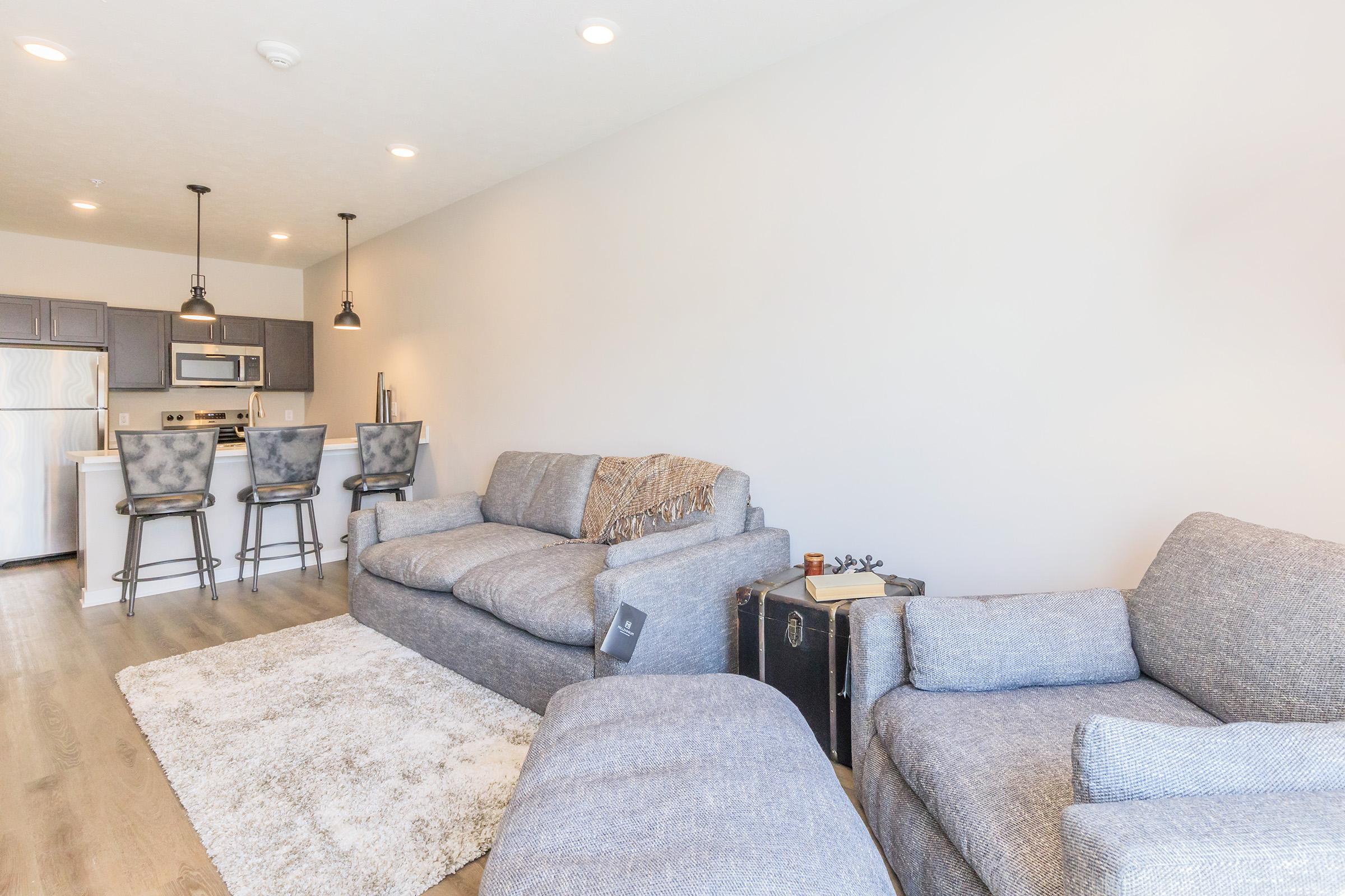 A cozy living room featuring a light gray sectional sofa, an armchair, and a plush area rug. The space includes a sleek kitchen counter with bar stools and modern appliances in the background. The walls are painted a soft neutral color, creating a bright and inviting atmosphere.