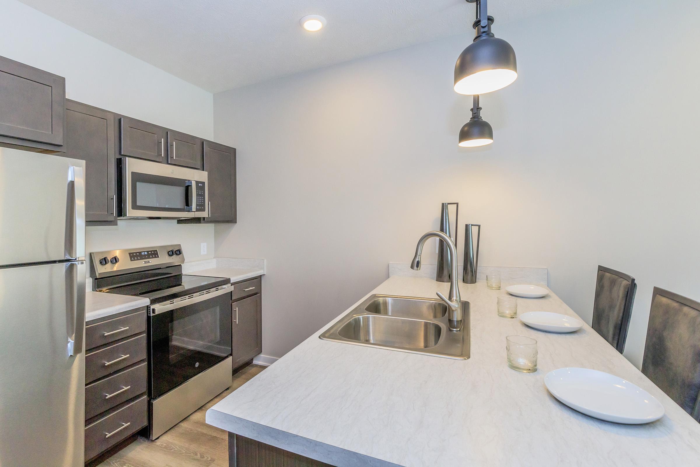 Modern kitchen featuring stainless steel appliances, including a refrigerator, microwave, and oven. A large island with a double sink is central, surrounded by four dining chairs. The cabinetry is dark wood, and there are two hanging pendant lights above the island, creating a bright and inviting atmosphere.