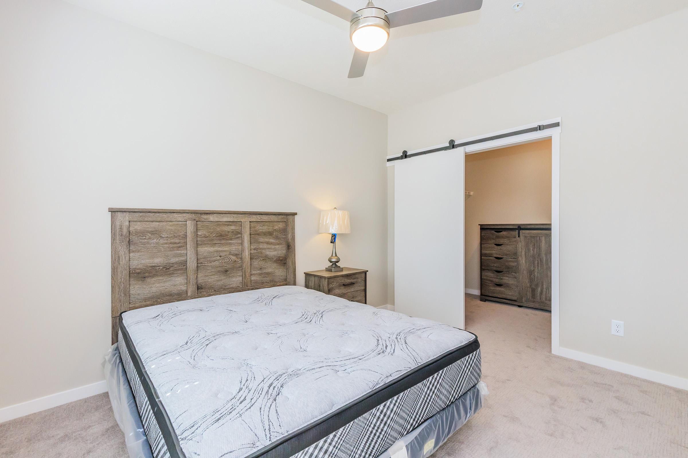 A simple bedroom featuring a bed with a patterned mattress, a bedside lamp on a wooden nightstand, and a sliding door leading to a closet. The walls are painted a light color, and the room has a ceiling fan. Natural light enhances the neutral tones and cozy atmosphere.