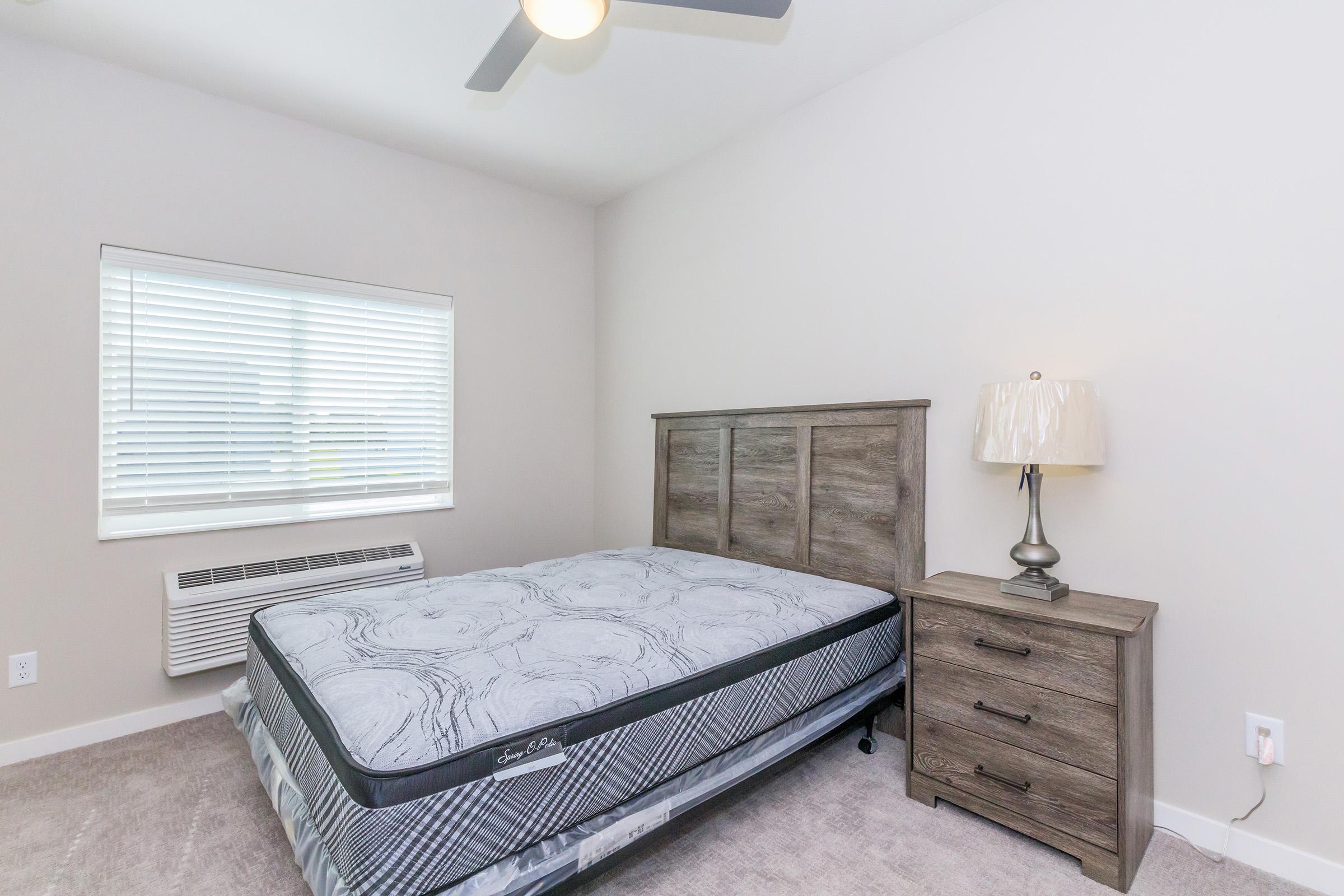 A simple, well-lit bedroom featuring a queen-sized bed with a gray mattress and a wooden headboard. There is a bedside table with a lamp next to the bed. A window with white blinds allows natural light to enter, and an air conditioning unit is situated below the window. The walls are painted in a light color, enhancing the spacious feel.