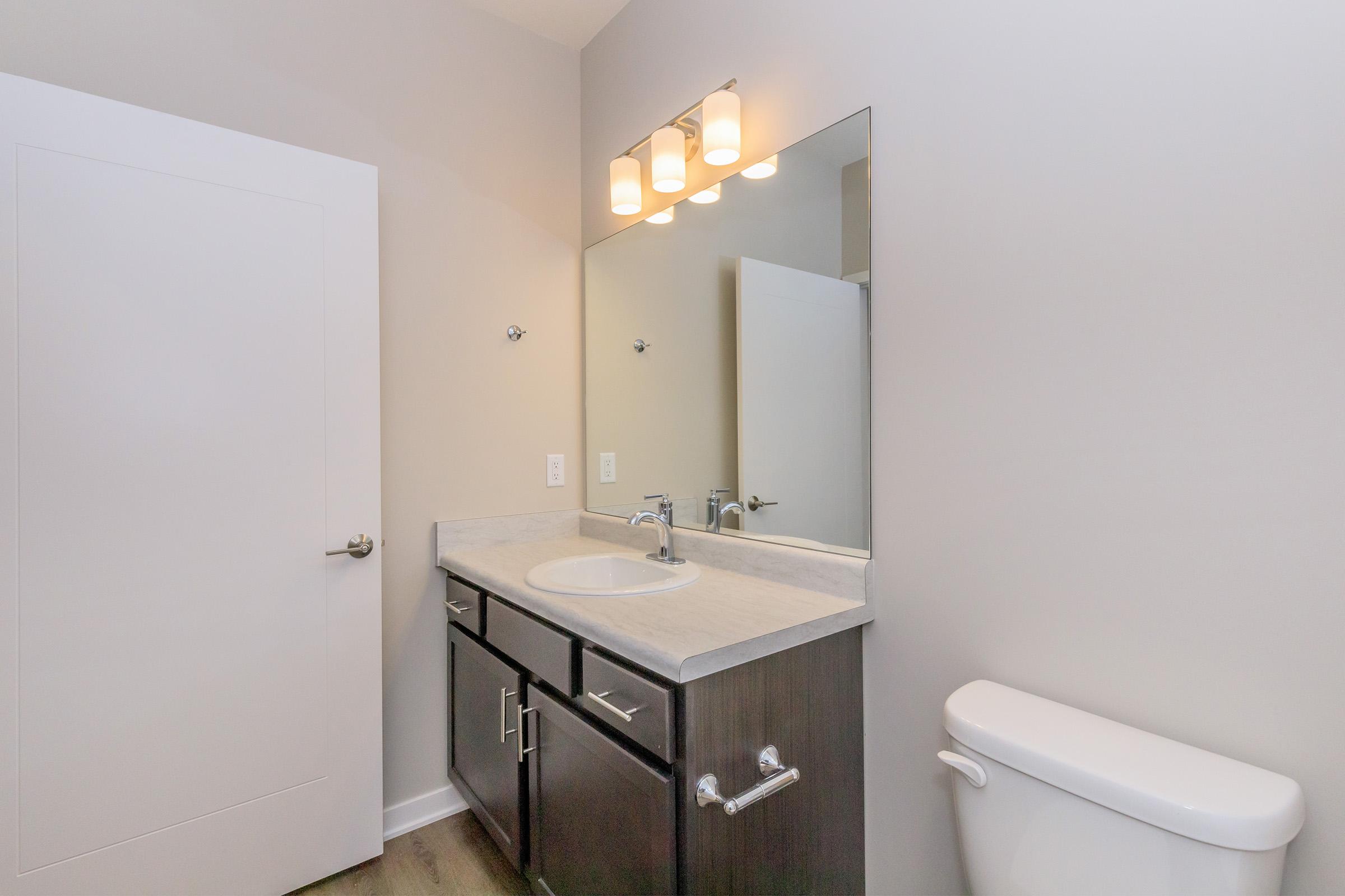 A modern bathroom featuring a white vanity with a sink and a countertop, a large mirror above the sink, and three light fixtures. There is a white toilet to the right and a light grey door on the left. The walls and floor have neutral tones, creating a clean and contemporary aesthetic.