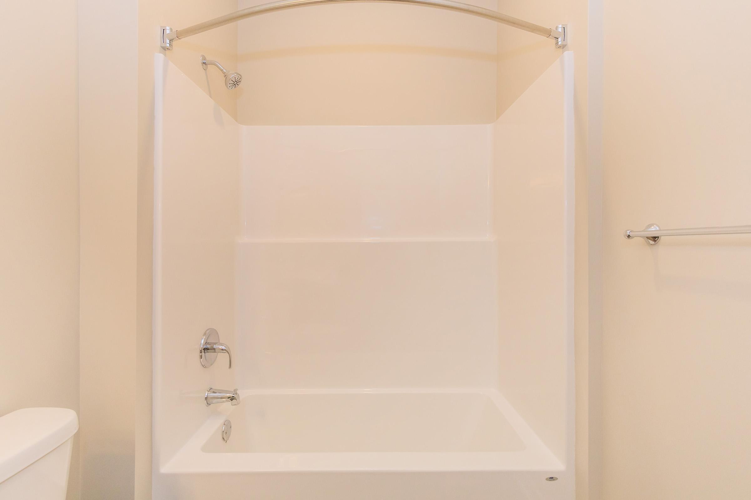 A clean, empty shower stall featuring a white tub and a chrome showerhead. The walls are smooth and white, and there is a towel bar on one side. A white toilet can also be seen partially in the frame. The overall setting is minimalistic and bright.