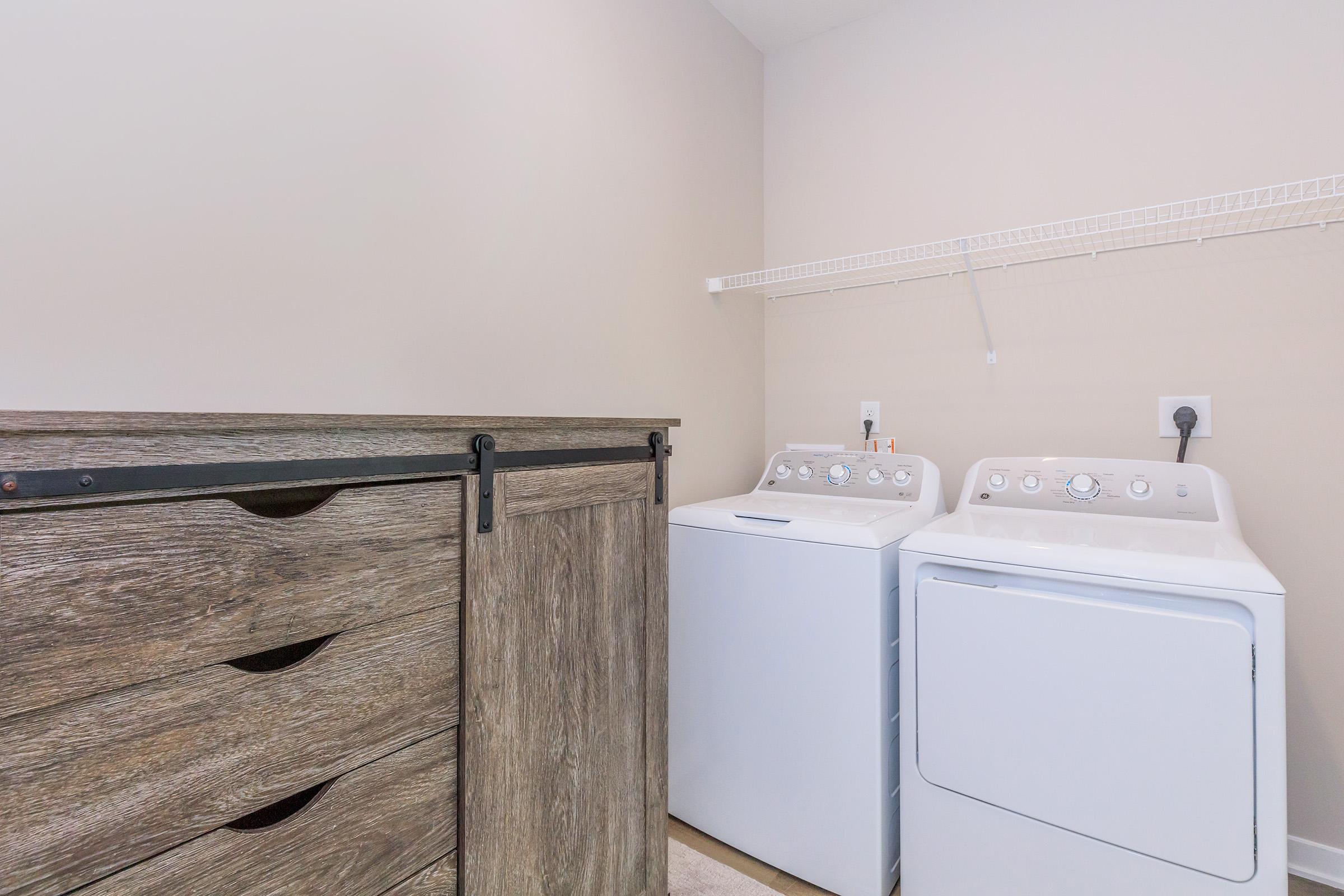 A laundry room featuring a washing machine and dryer side by side, with a wooden storage unit nearby. The walls are painted in a neutral color, and there is a wall-mounted shelf above the appliances for additional storage. The floor is light-colored carpet or tile.