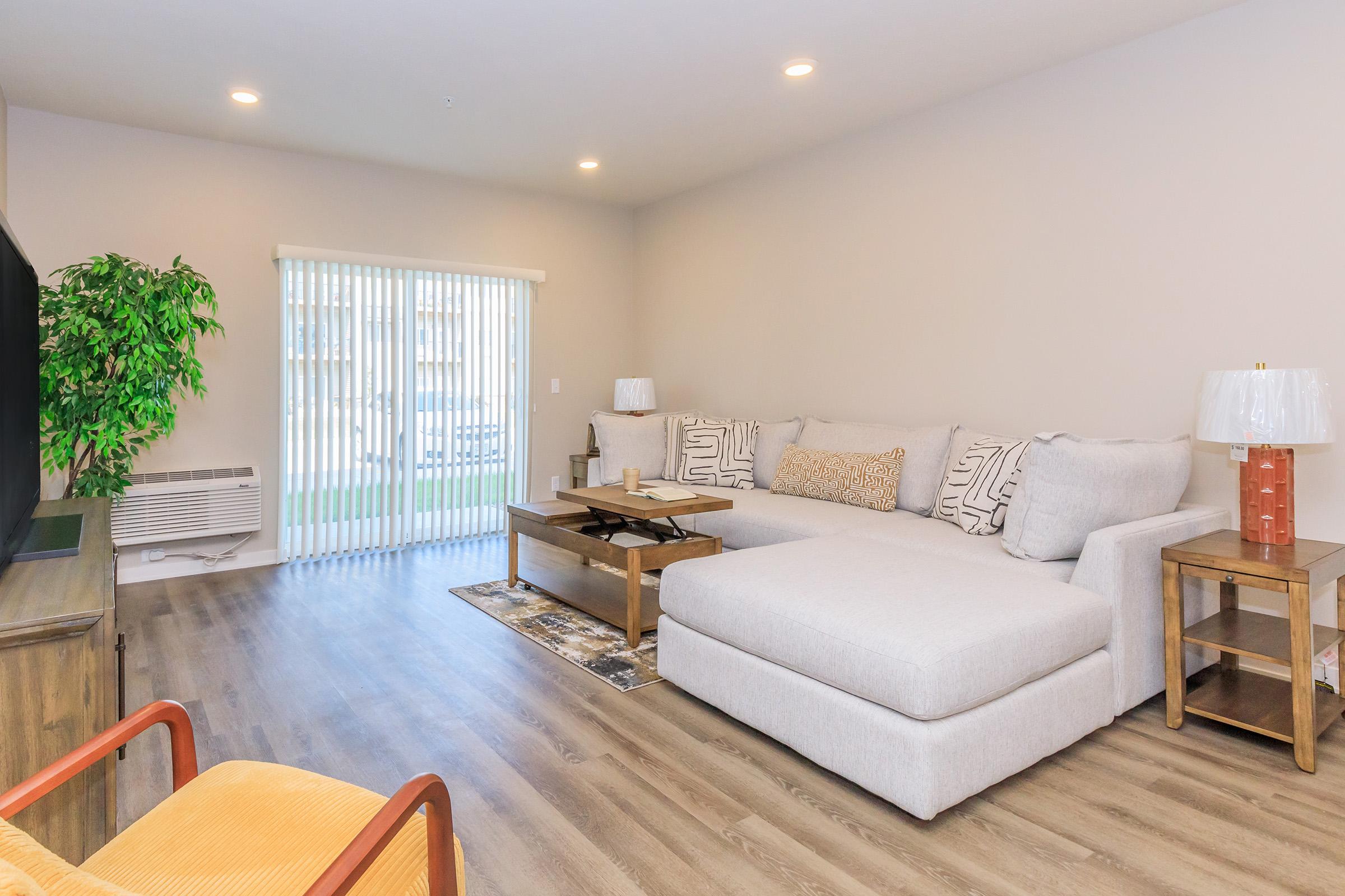 A modern living room featuring a large sectional sofa with decorative pillows, a wooden coffee table, and an orange accent chair. There's a TV on a wooden console and large sliding doors with vertical blinds leading to an outdoor space. A potted plant adds a touch of greenery to the light-colored walls and flooring.